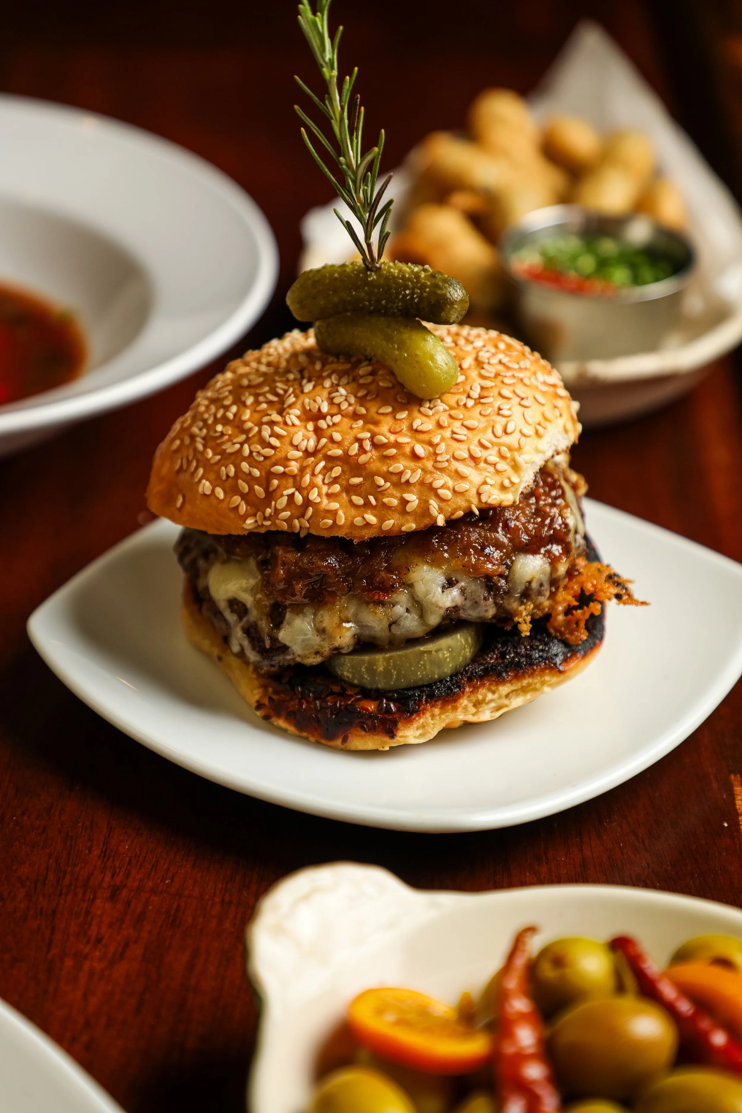 Close-up of a gourmet burger with sesame seed bun, pickles, melted cheese, and a sprig of rosemary and pickles on top, served on a white plate with side dishes in the background.