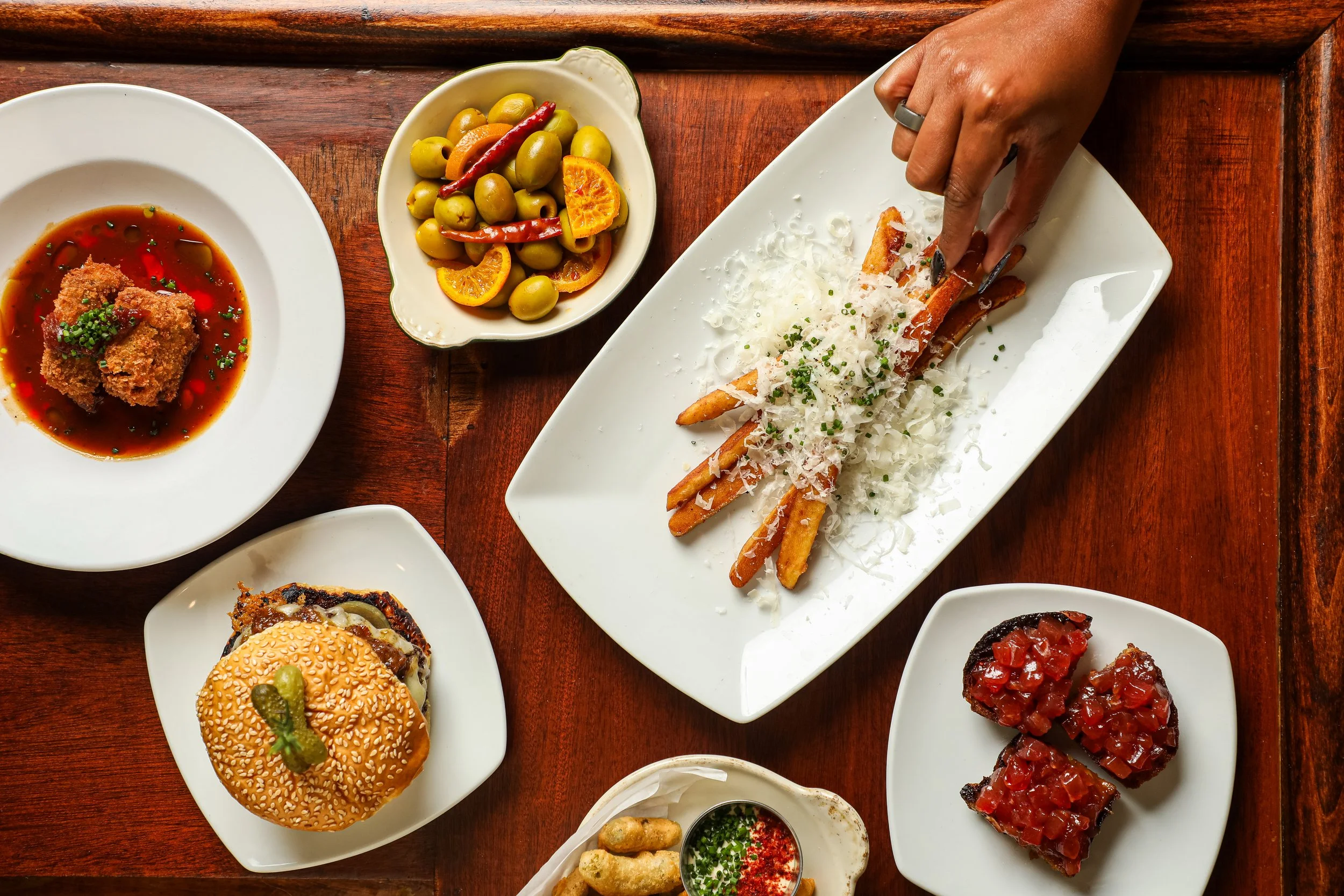 Assorted dishes of french cuisine on white plates and bowls on a wooden table, including spring rolls, green olives with orange slices and red chili, fried meat with sauce, and a cheeseburger with pickle.