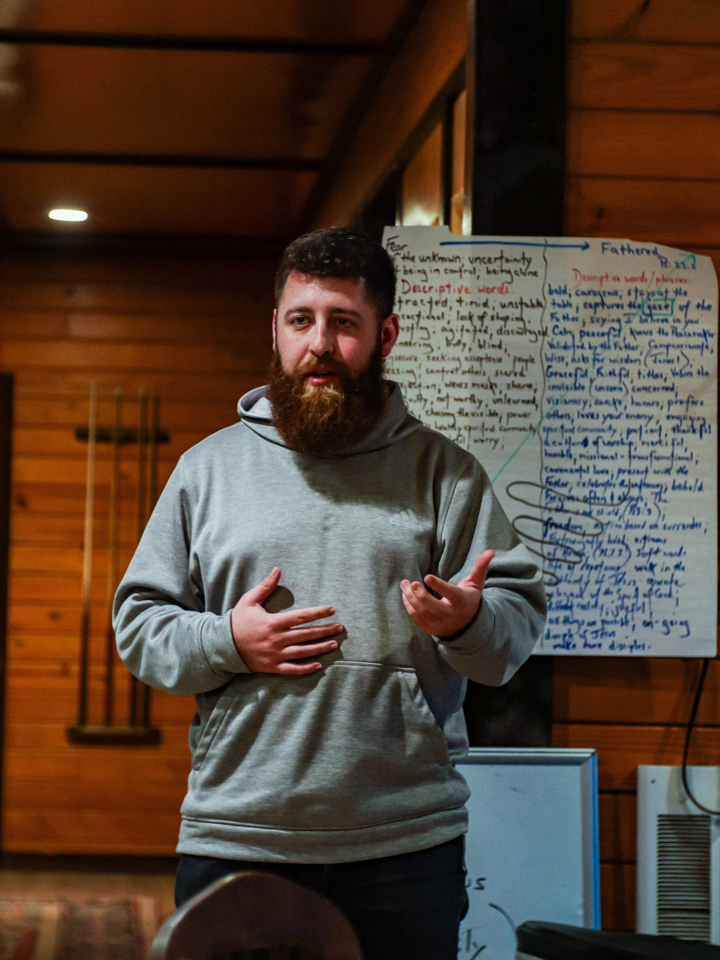A man with a beard wearing a grey hoodie giving a presentation in front of a whiteboard filled with handwritten notes and diagrams.