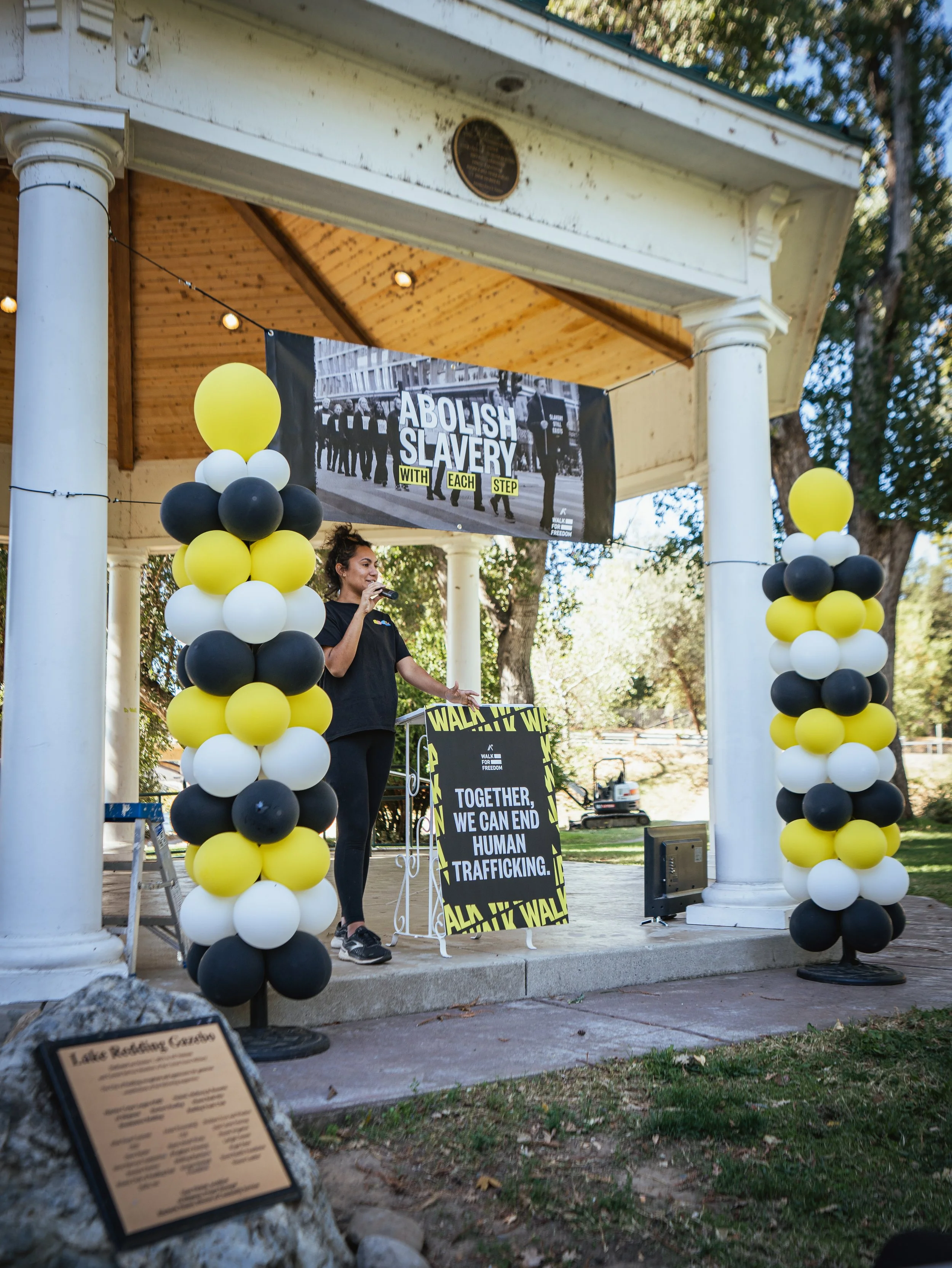 Woman speaking at a rally with signs advocating to abolish slavery and human trafficking, decorated with black, white, and yellow balloons in a park gazebo.