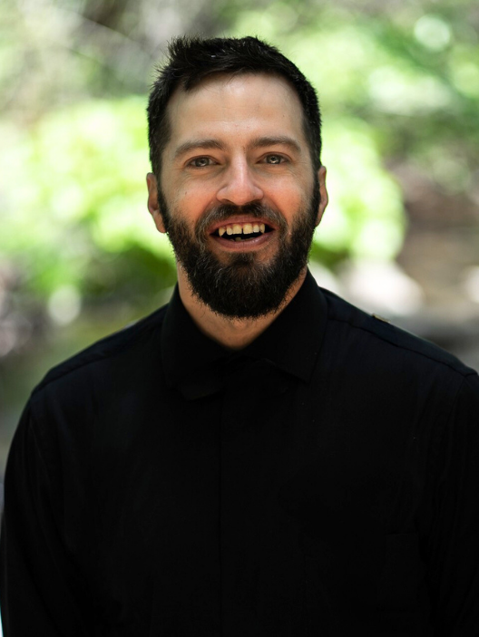 A smiling man with a beard and short dark hair, wearing a black shirt, outdoors with a blurred green background.