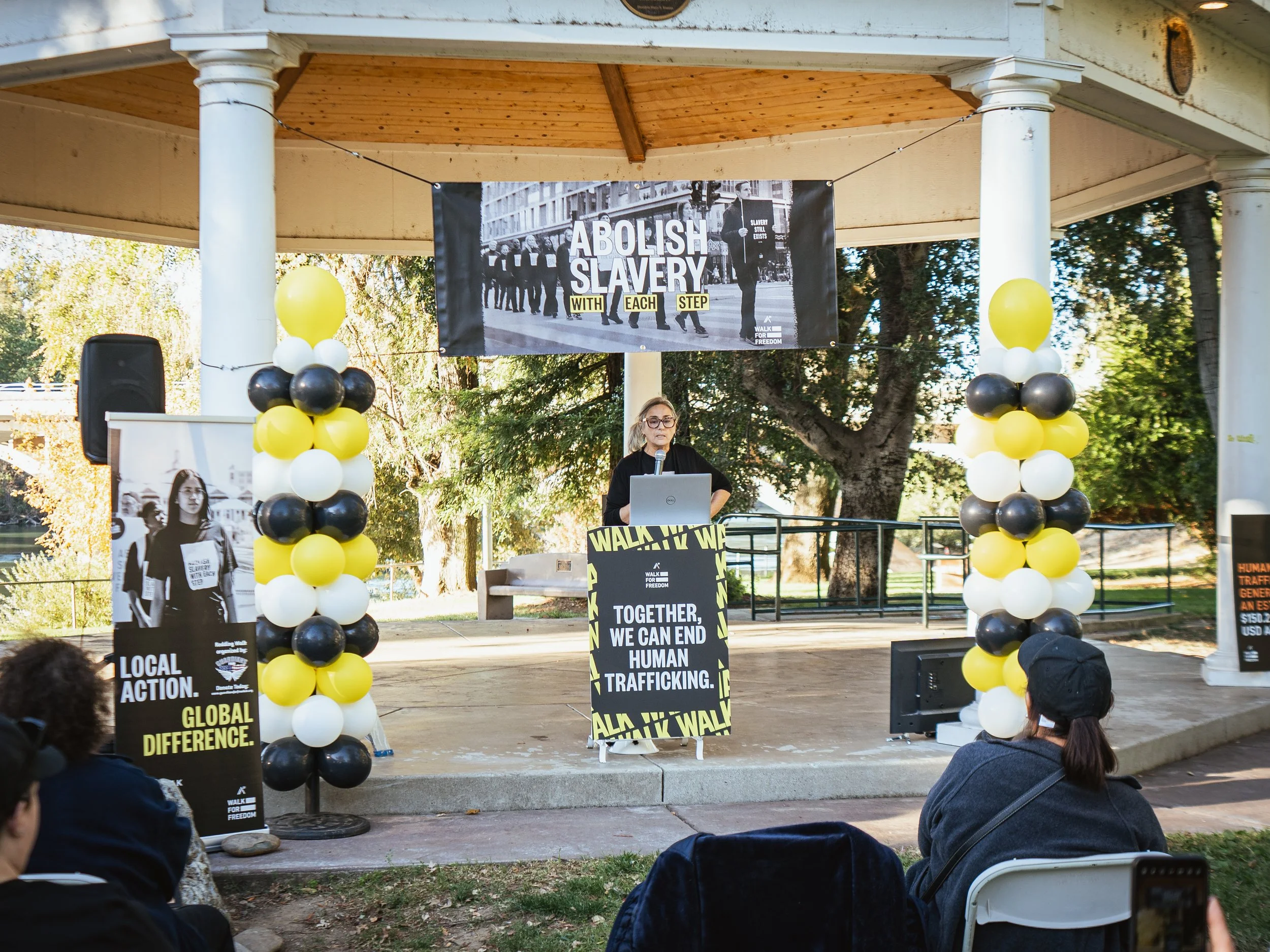 A woman speaking at a public event about ending human trafficking, outdoors under a pavilion, with black, yellow, and white balloons, posters, and a banner that reads 'Abolish Slavery with Each Step' and 'Together, We Can End Human Trafficking.'