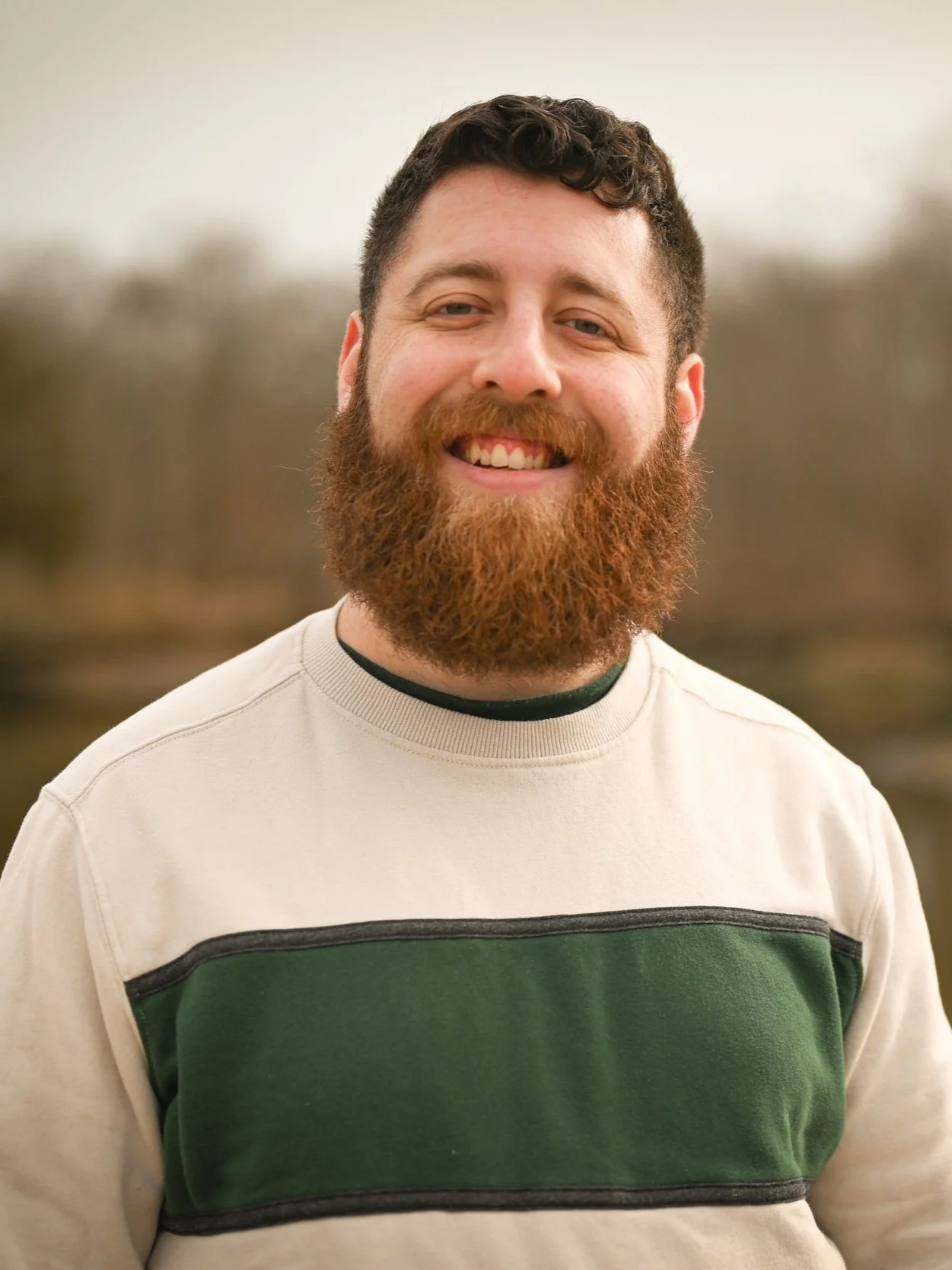 A smiling man with a reddish beard and short dark hair outdoors, wearing a beige sweatshirt with a green and black stripe across the chest.