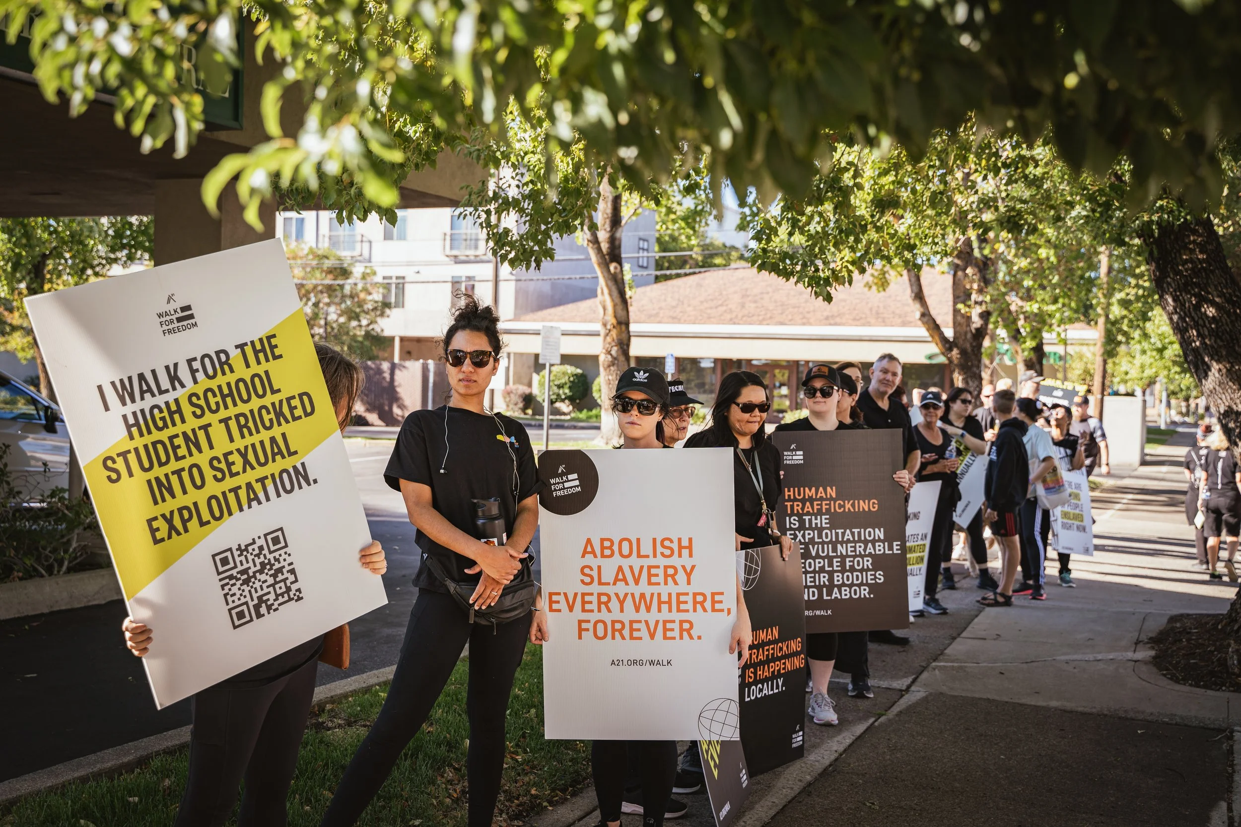 A group of protesters holding signs about human trafficking, slavery, and sexual exploitation during a demonstration on a tree-lined sidewalk.