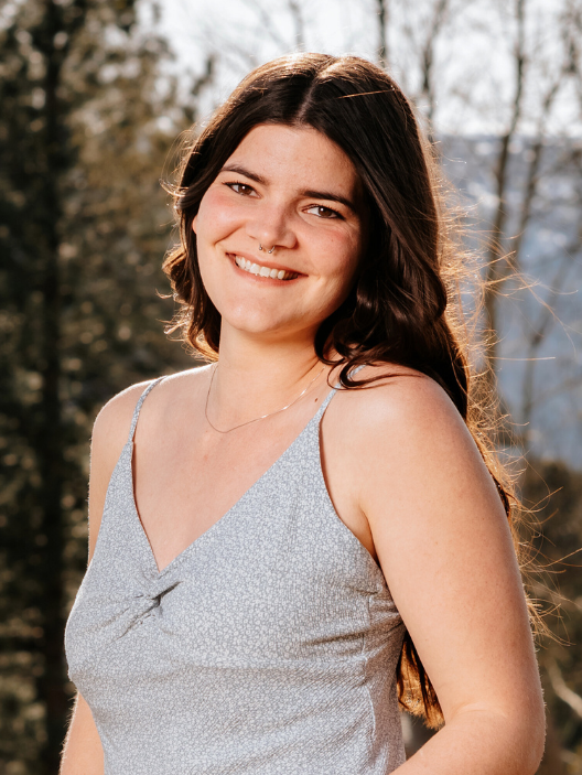 Smiling young woman with long dark hair wearing a light blue spaghetti strap top outdoors with trees and mountains in the background.