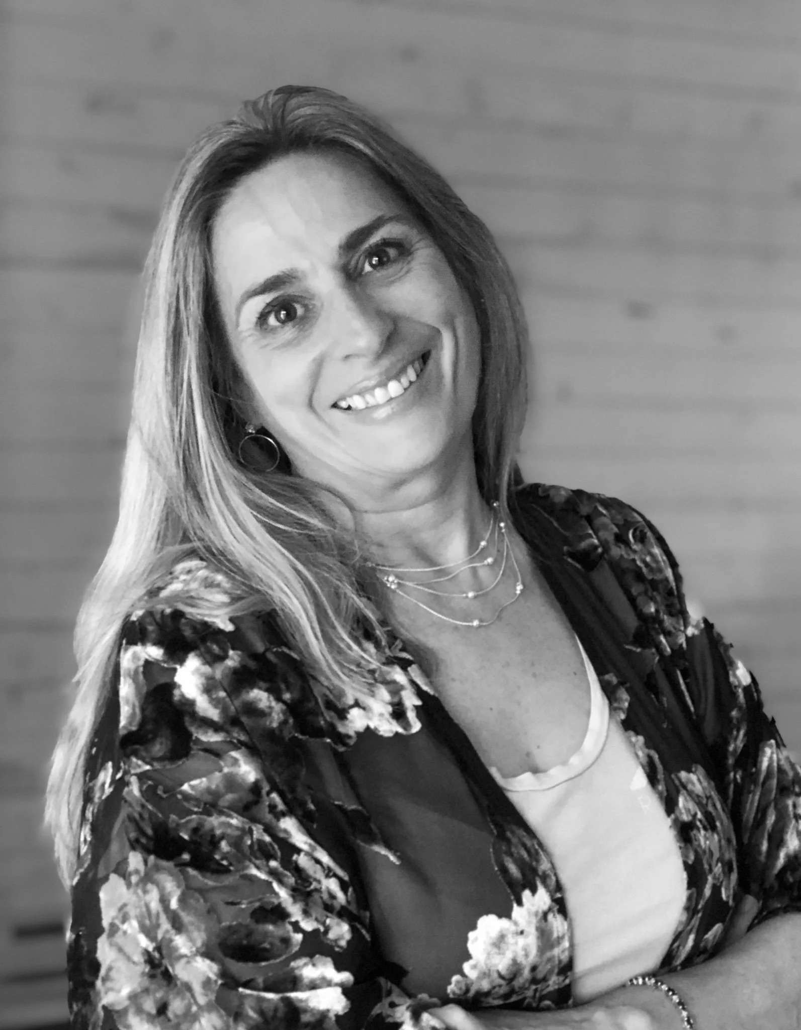 A smiling woman with blonde hair, wearing a blue top, jewelry including earrings and a necklace, and posing against a plain background.