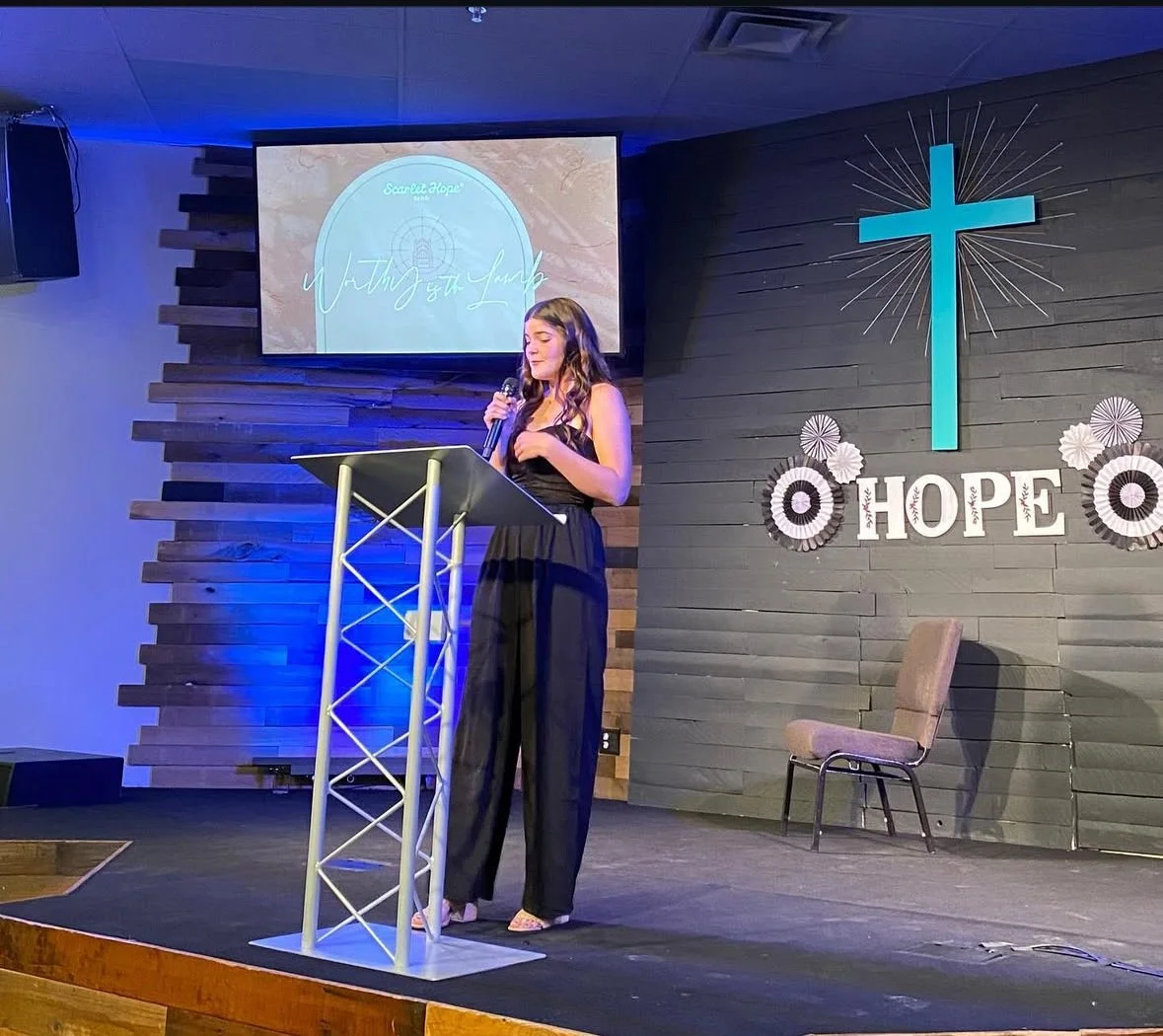 A young woman in a black dress standing at a podium, speaking into a microphone in a church or religious setting. Behind her, a large screen displays the text 'Walking in Faith'. The wall behind her has a large blue cross and decorative elements with the word 'HOPE'.
