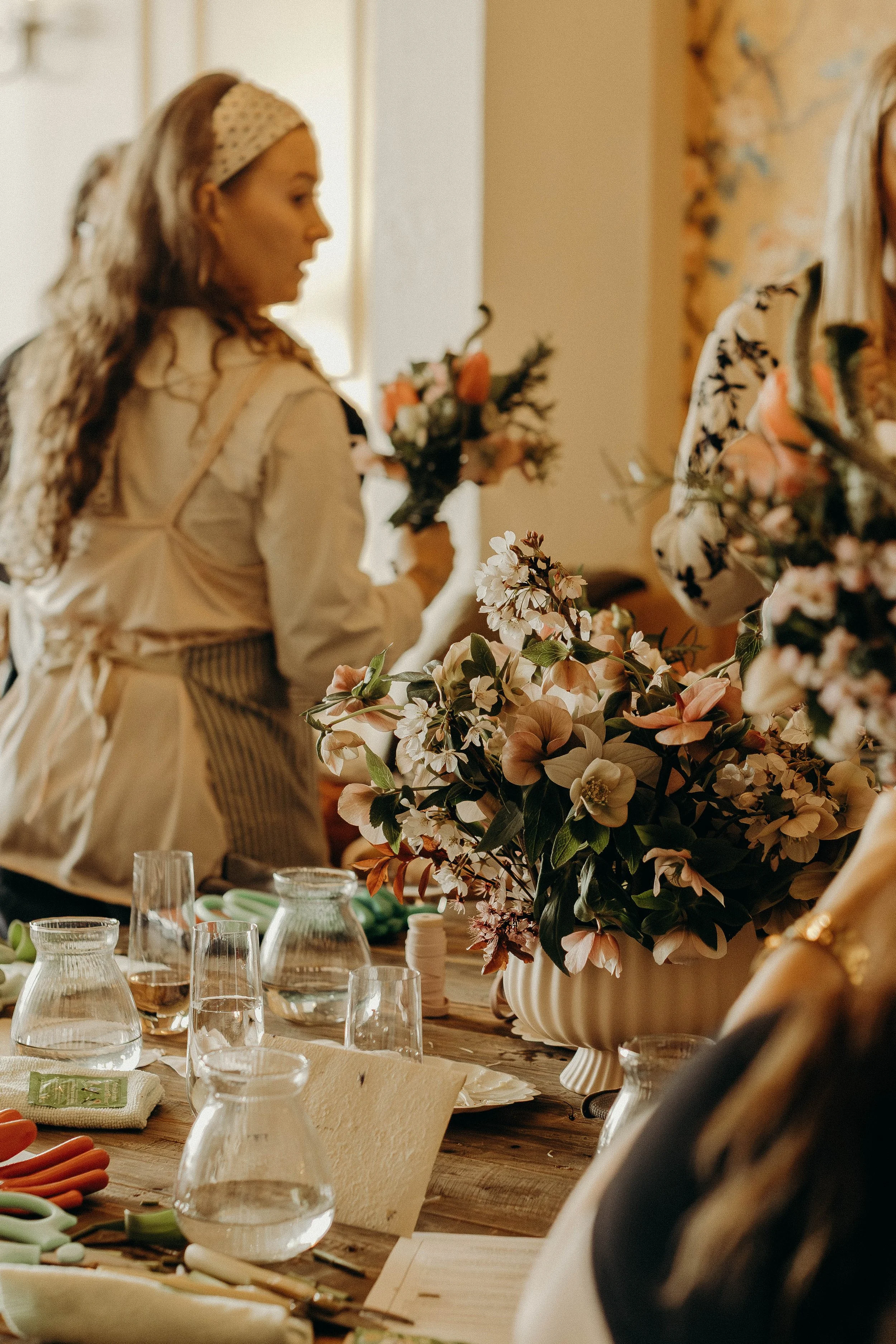 A woman in a beige apron and polka dot headband arranging flowers on a table, with glass water pitchers and floral arrangements in the foreground., captured by Alison May Photography.