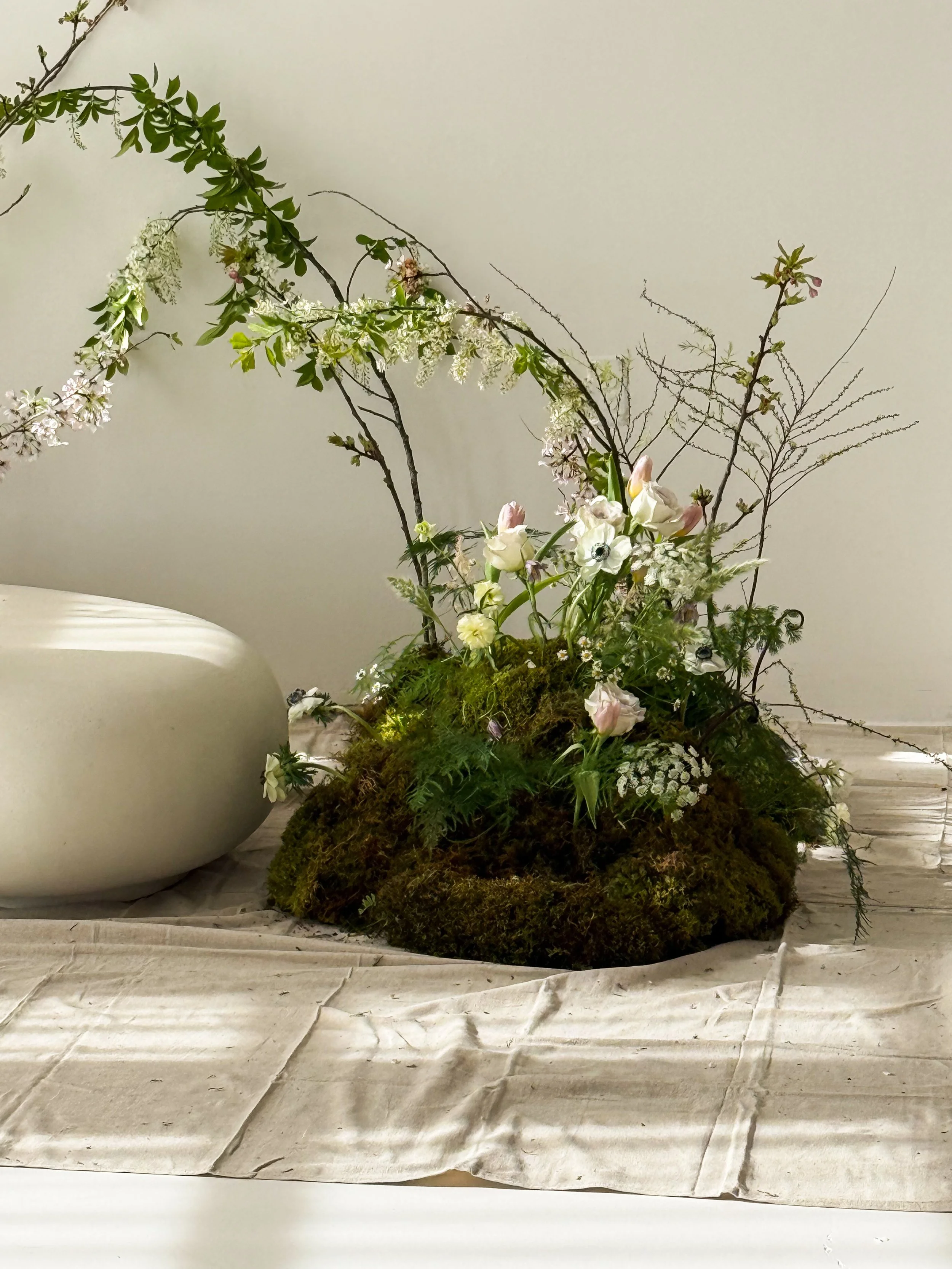 Elegant flower arrangement with white and pink flowers, green foliage, and moss, set on a white table with a white background.