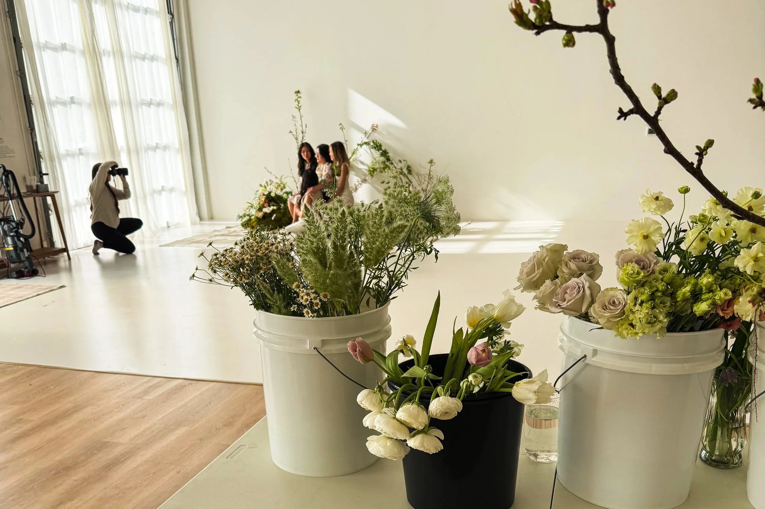 A woman takes a photo of three women sitting on a white backdrop decorated with flowers in a bright room with large windows and sheer curtains. In the foreground, there are buckets filled with various flowers and a glass of water.