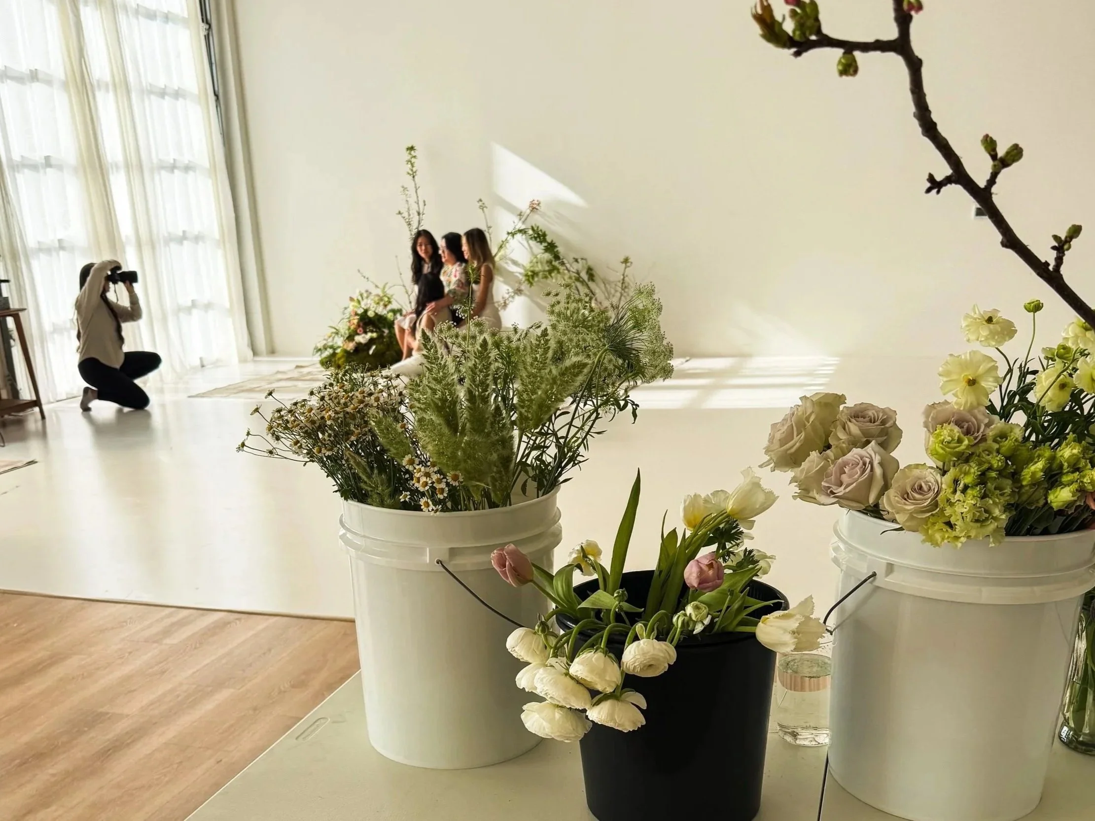 Three women sitting on a bench in front of a plain wall, with a photographer kneeling nearby taking their picture. In the foreground, there are three pots of flowers, with the one in the center containing white tulips and a glass of water.