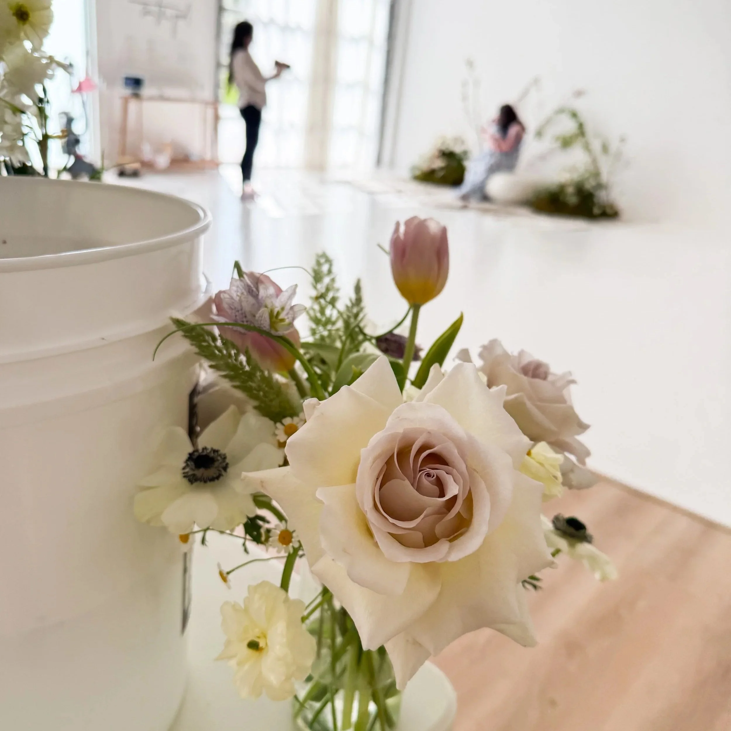 Close-up of a small bouquet of pastel flowers including a large pale pink rose, white daisies, and pink tulips, placed near a white container in a bright, modern room with a blurred background of two women, one sitting and one standing.