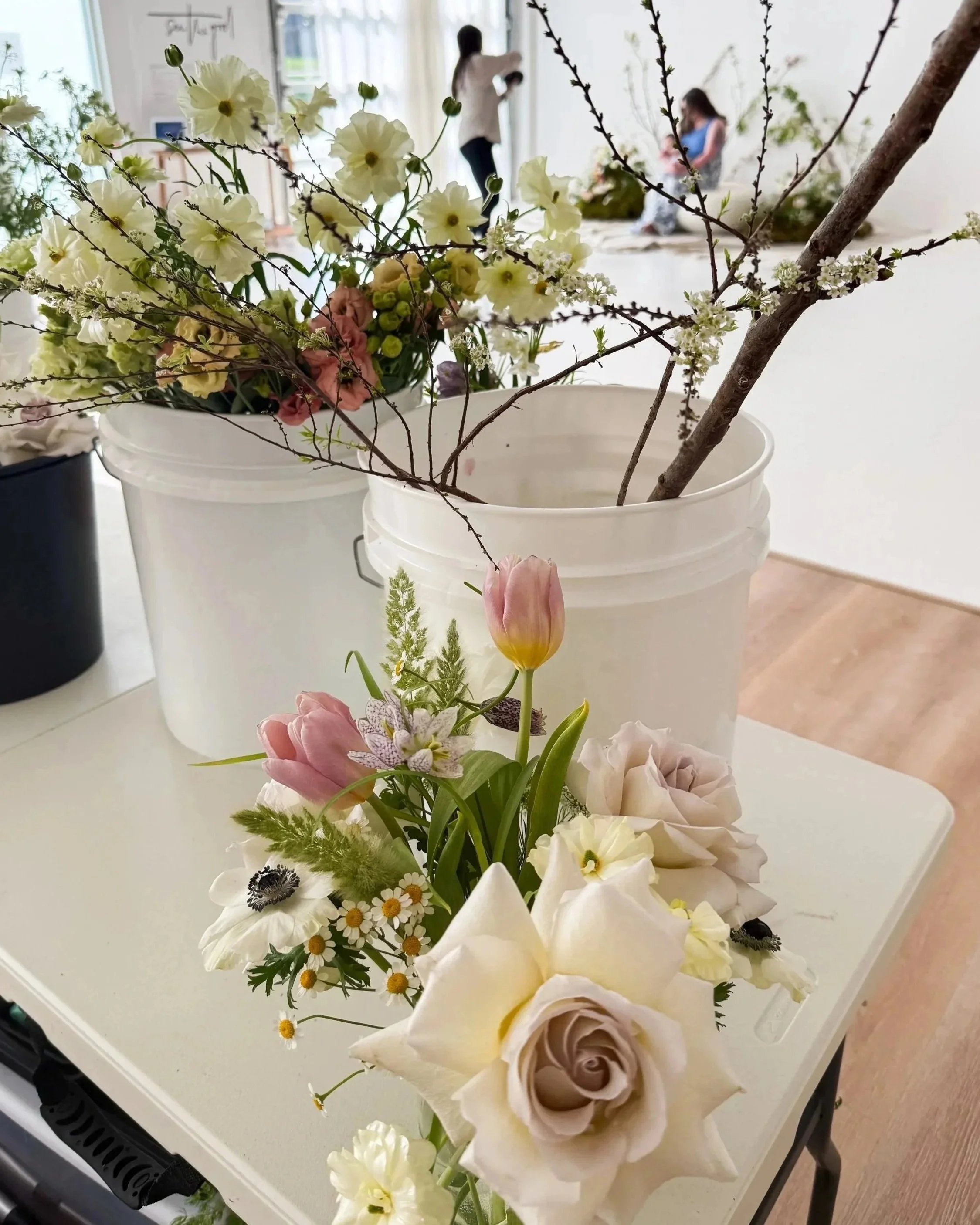 A small flower arrangement with roses, tulips, daisies, and other flowers on a white table, with a large white bucket containing branches and flowers in the background.