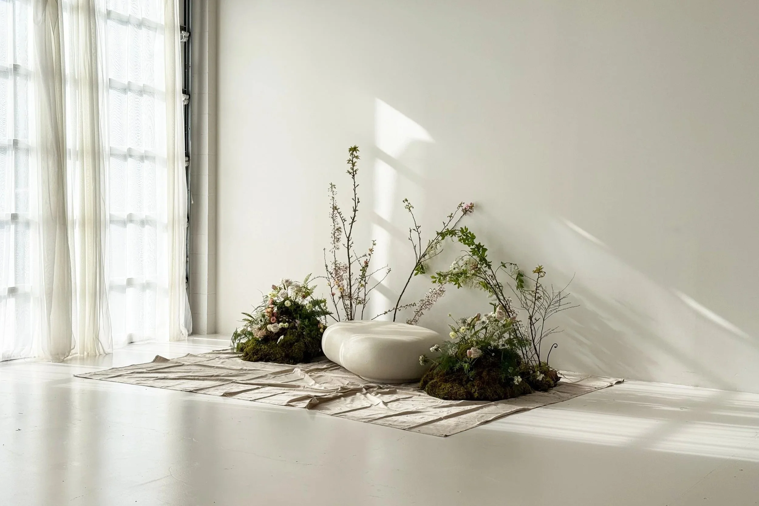 Minimalist interior with white walls and floors, sheer curtains letting in natural light, a fabric-covered base with floral arrangements and twigs, and a large white rounded vessel.