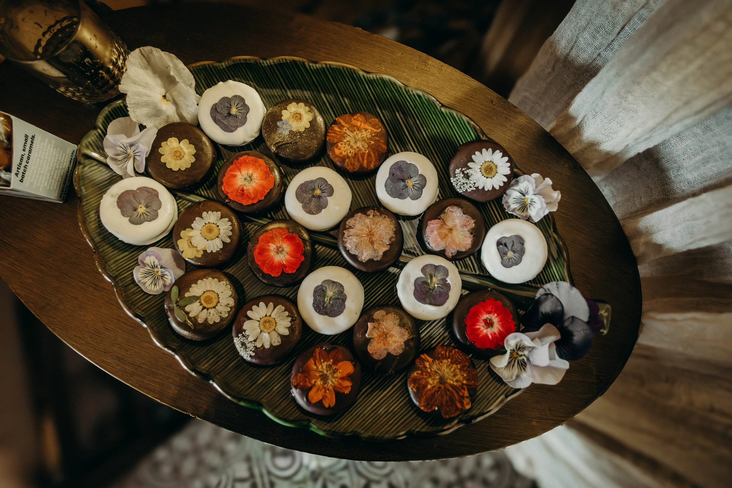 A decorative tray with chocolate and white confections topped with edible flower decorations, placed on a wooden surface with scattered edible flowers around it, made by Jenn Hughes and captured by Alison May Photography.