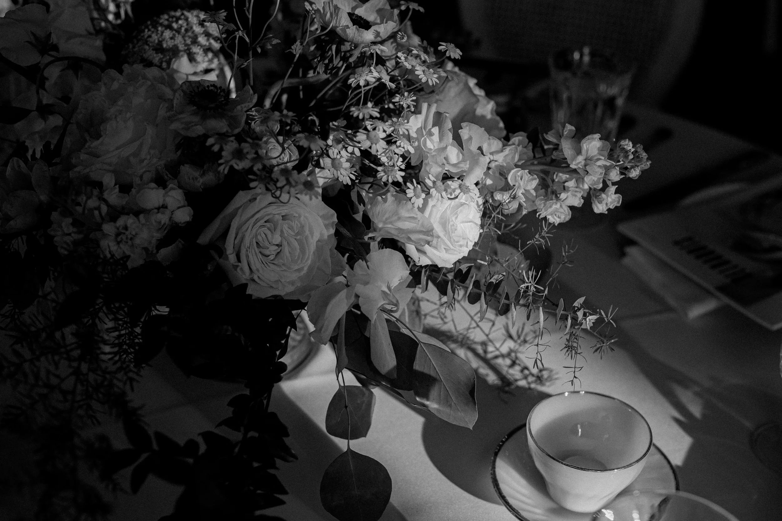 Black and white photograph of a floral centerpiece with roses and other flowers on a table, along with a candle in a glass holder, and a glass of water in the background., captured by Kay Gilgan.