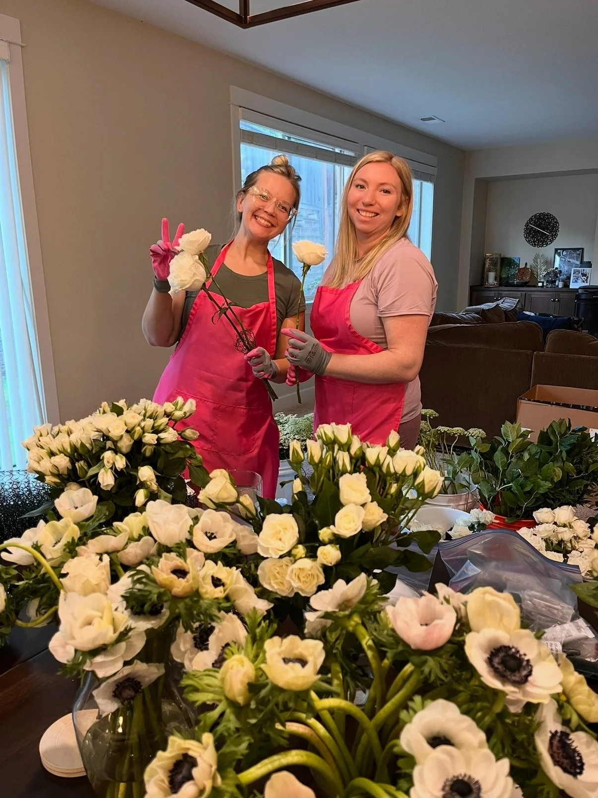 Two smiling guests in bright aprons holding fresh flowers during a Bloom Bar workshop, surrounded by white anemones and greenery.