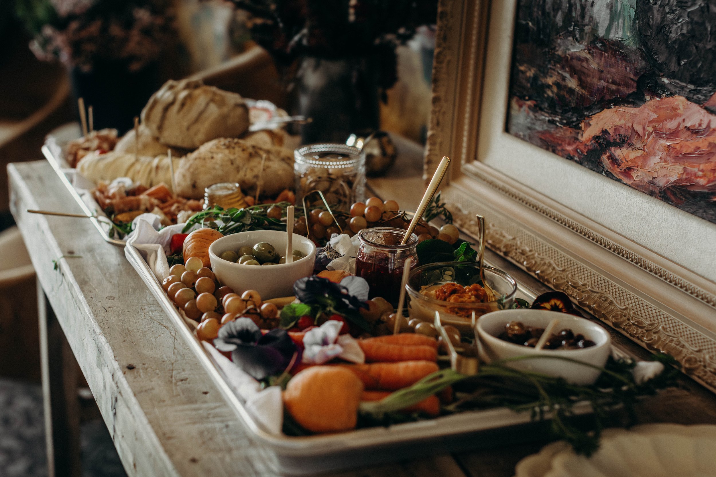 A tray with various appetizers including bread, grapes, carrots, olives, and jams set on a rustic table, with a decorative frame and abstract painting in the background.