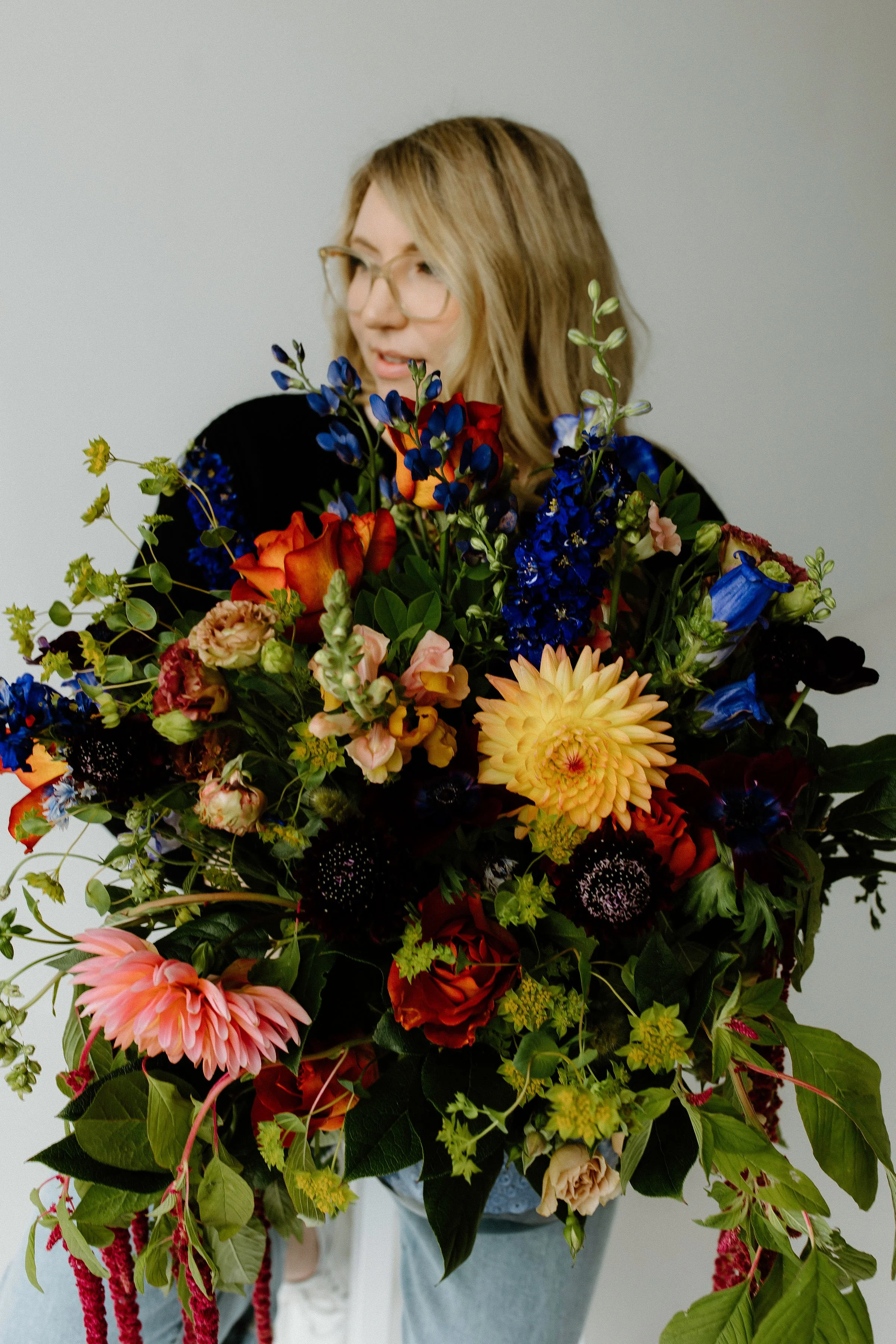 Founder holding an abundant floral arrangement with bold textures and saturated tones, featuring cobalt delphinium, rust mums, golden dahlias, and trailing amaranthus.