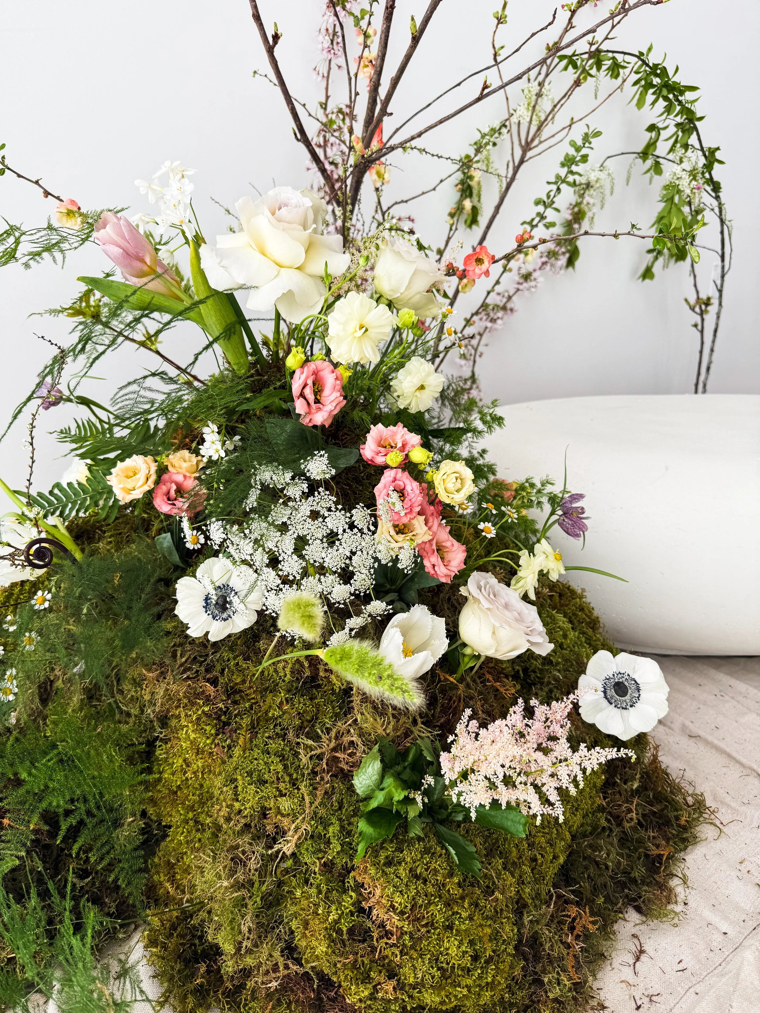 A floral arrangement with various white, pink, and yellow flowers, green ferns, and moss, placed on a white surface against a plain light background.