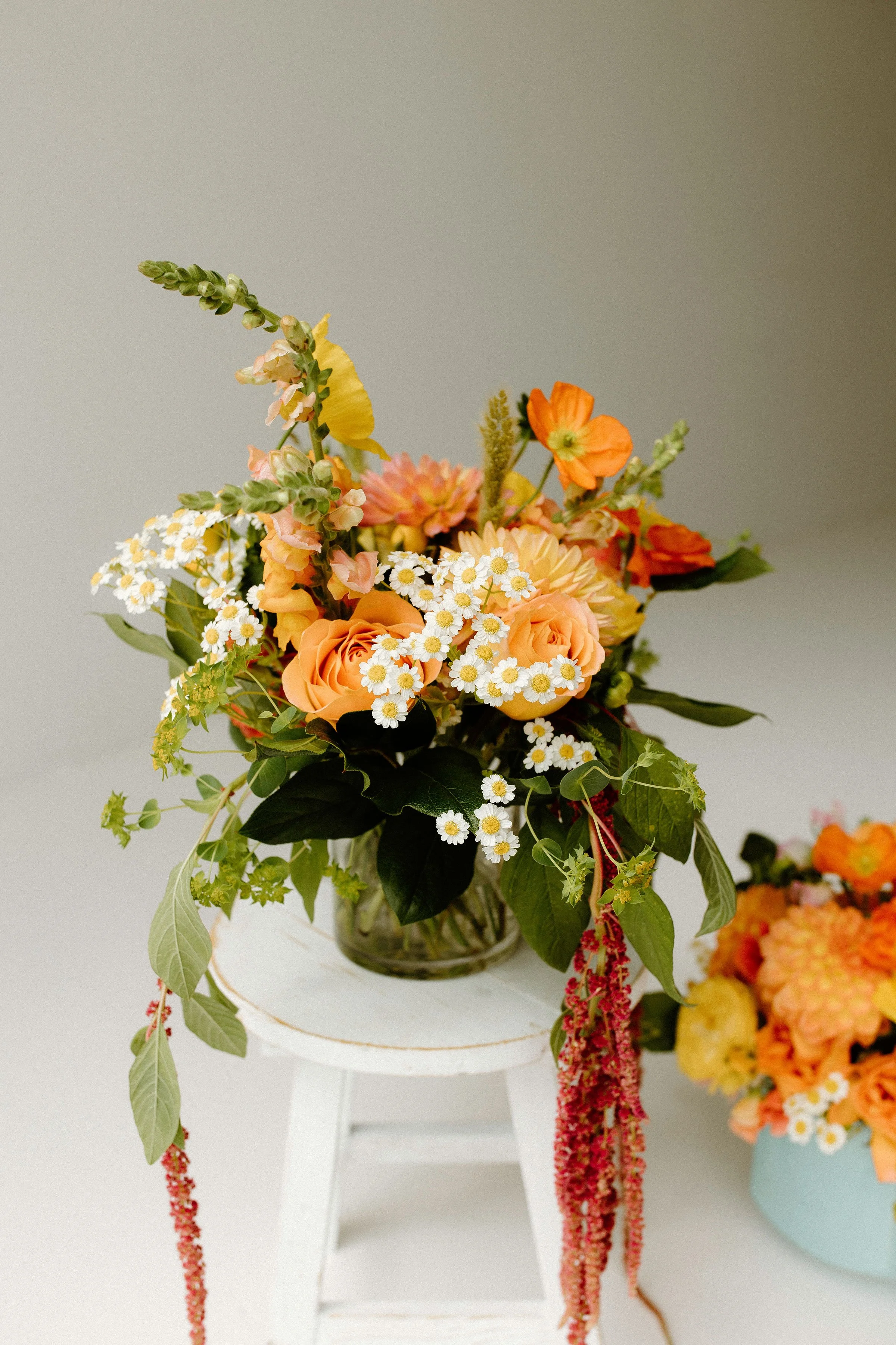 Lush Golden Hour bouquet with rich marigolds, creamy florals, pink garden roses, and trailing greenery, arranged in a full-bodied sculptural style in a clear glass vase
