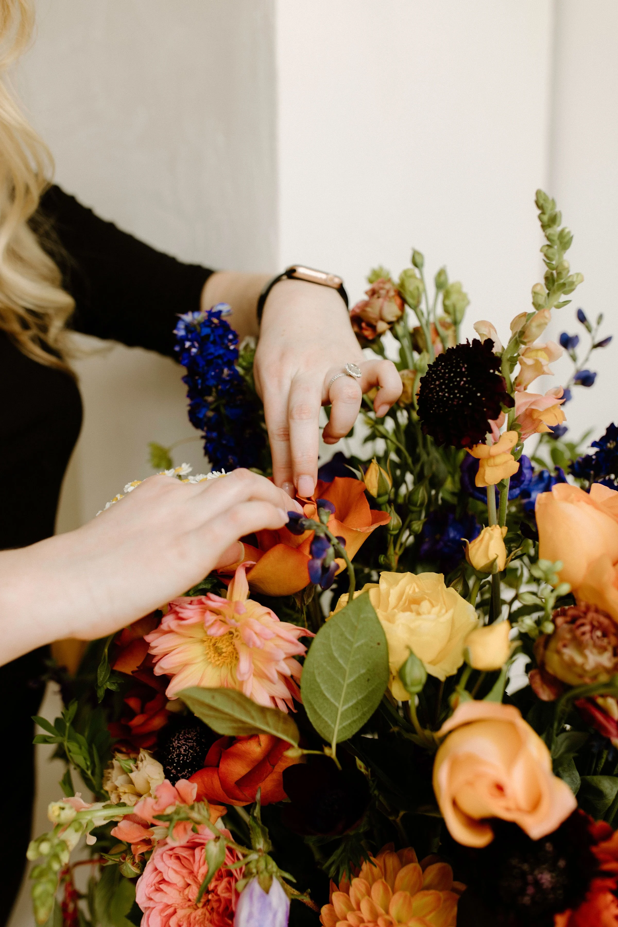 Close-up of founder arranging flowers for a private event centerpiece, featuring moody florals like navy delphinium, apricot snapdragons, orange poppies, and pale peach roses.