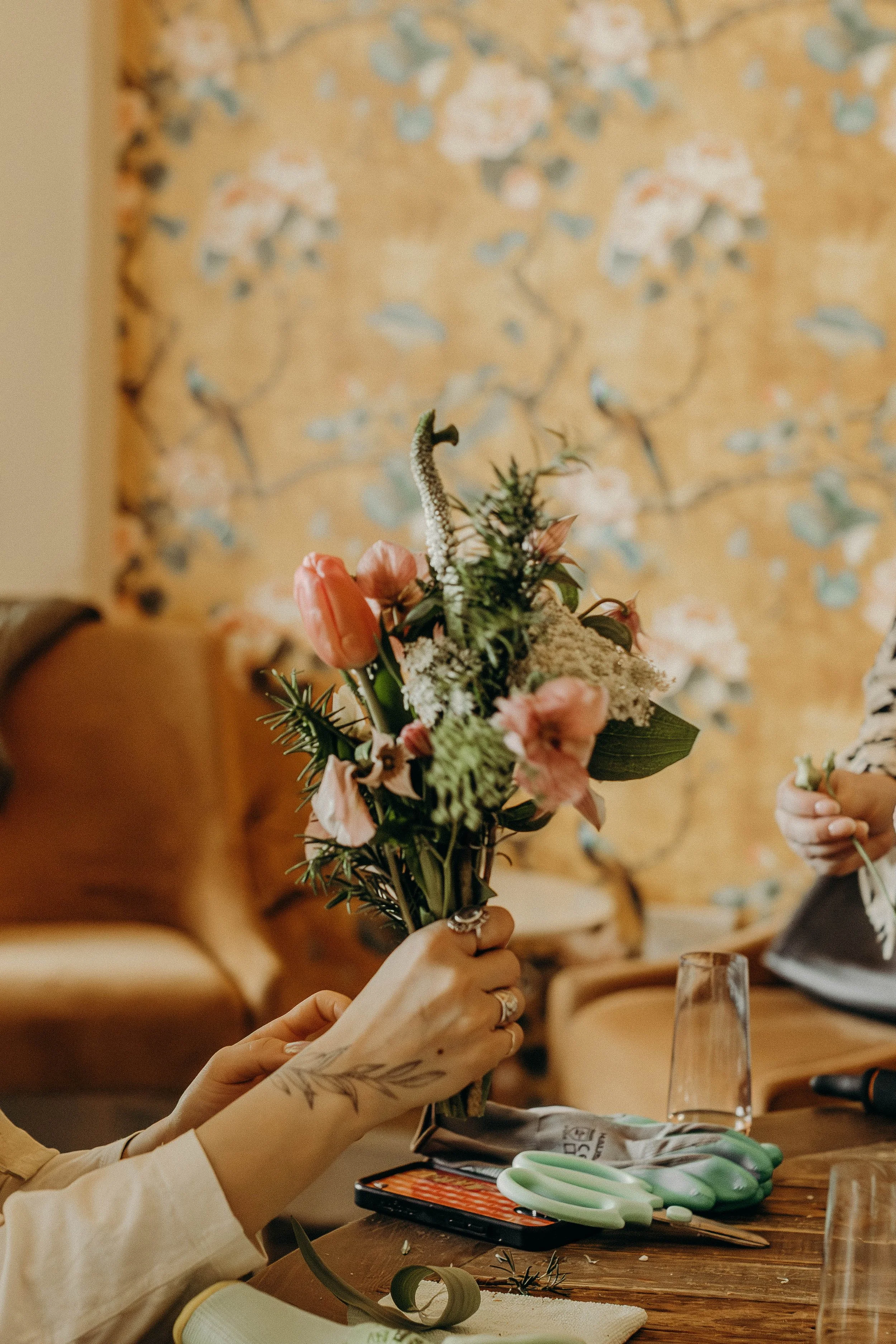 Person holding a bouquet of pink and green flowers, with a tattooed arm and a tattooed hand, on a wooden table with scissors and a smartphone nearby, in a room with floral wallpaper.