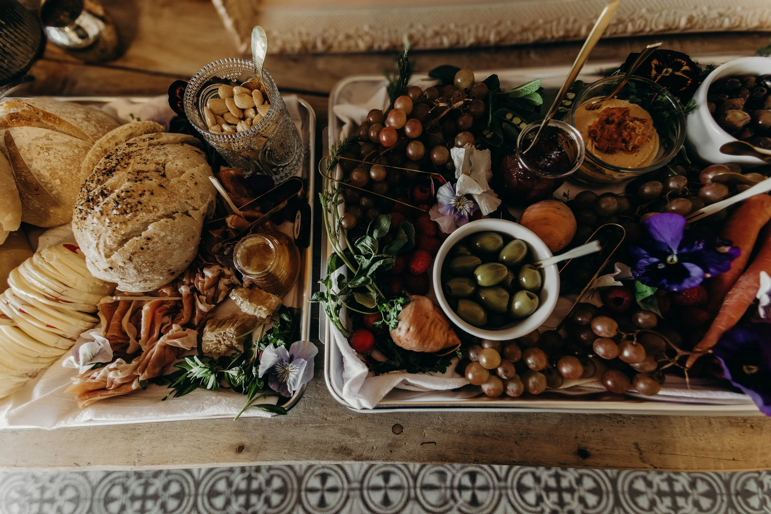 A breakfast spread with bread, cheese, nuts, preserves, grapes, olives, strawberries, flowers, and vegetables on a wooden table.