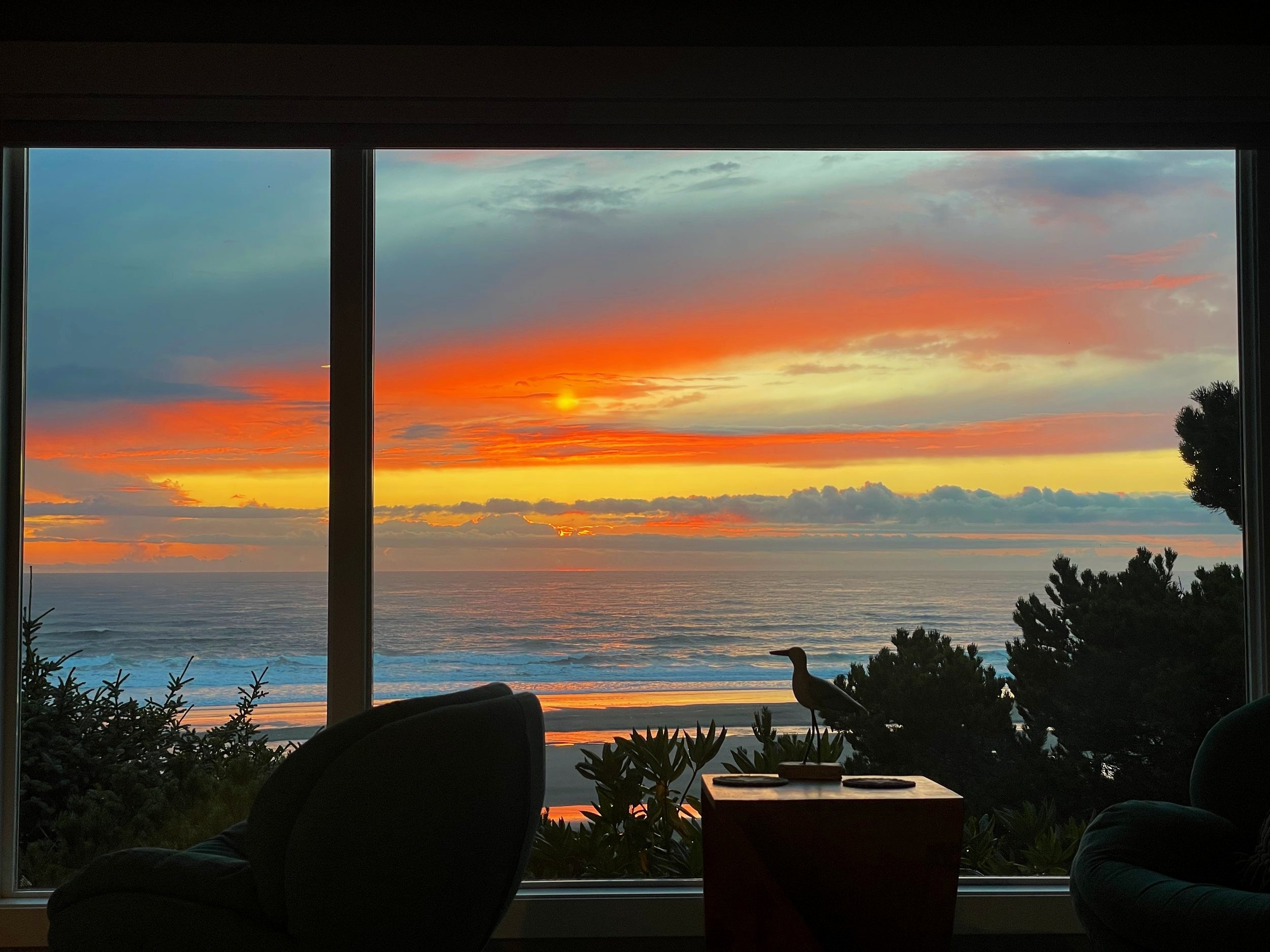 Golden hour over the Oregon Coast, viewed from indoors through a window, with a silhouetted seagull and glowing sky in warm orange, gold, and lavender tones.