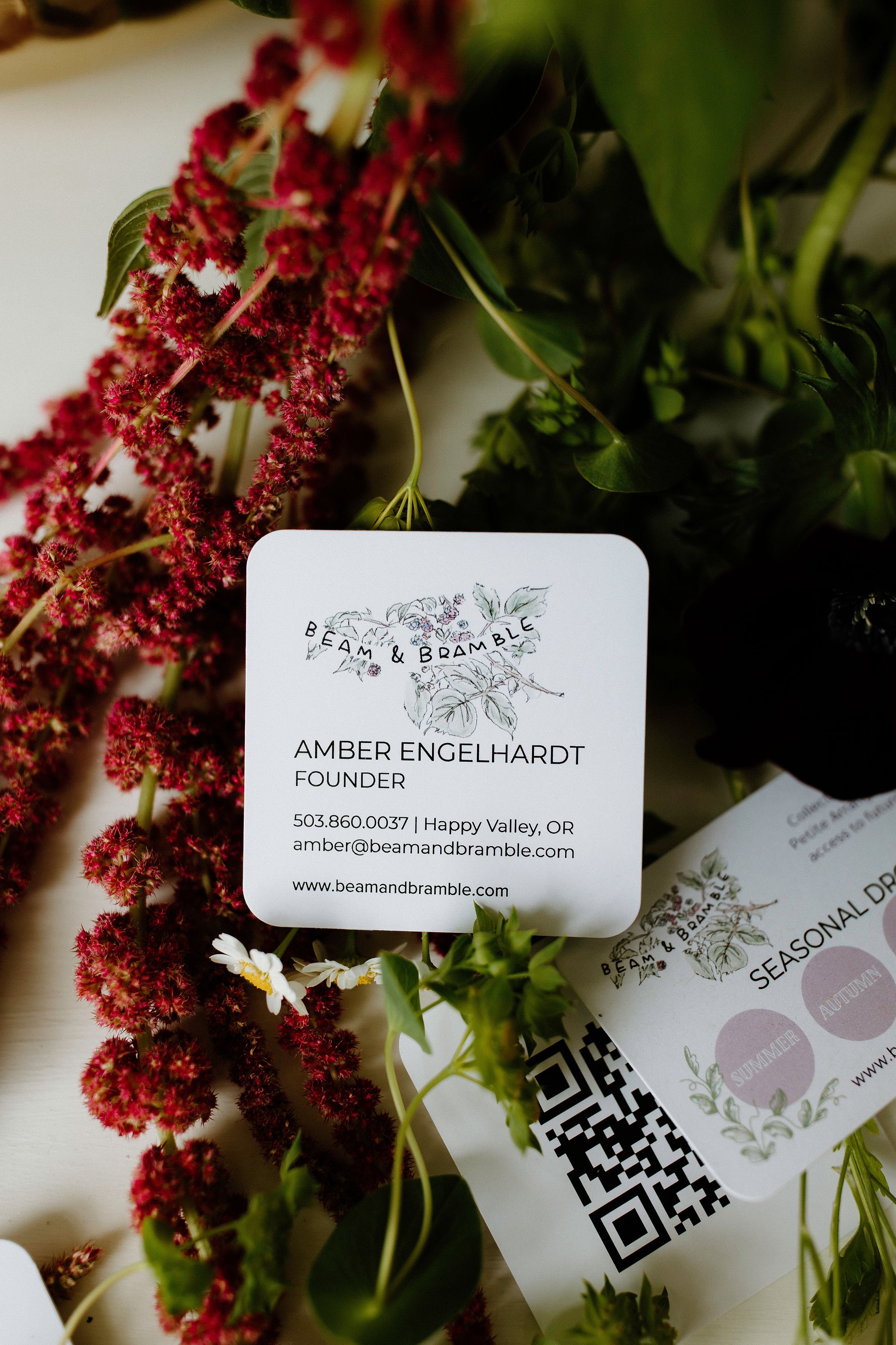Close-up of a Beam & Bramble business card featuring floral illustrations and contact info for founder Amber Engelhardt, surrounded by fresh flowers and greenery with a QR code partially visible.