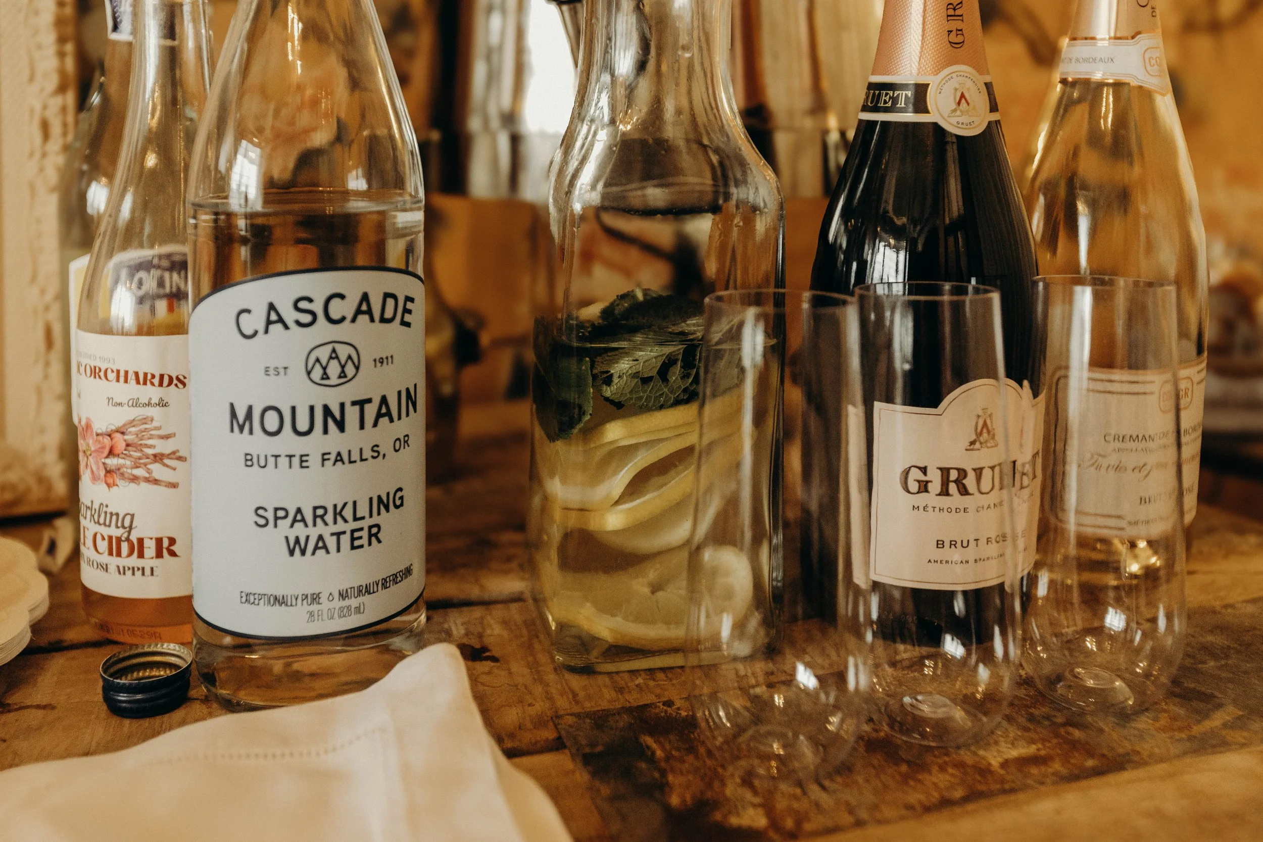 Various bottles and glasses on a wooden table, including sparkling water, apple cider, and a bottle of sparkling wine, with lemon and mint inside a glass pitcher.