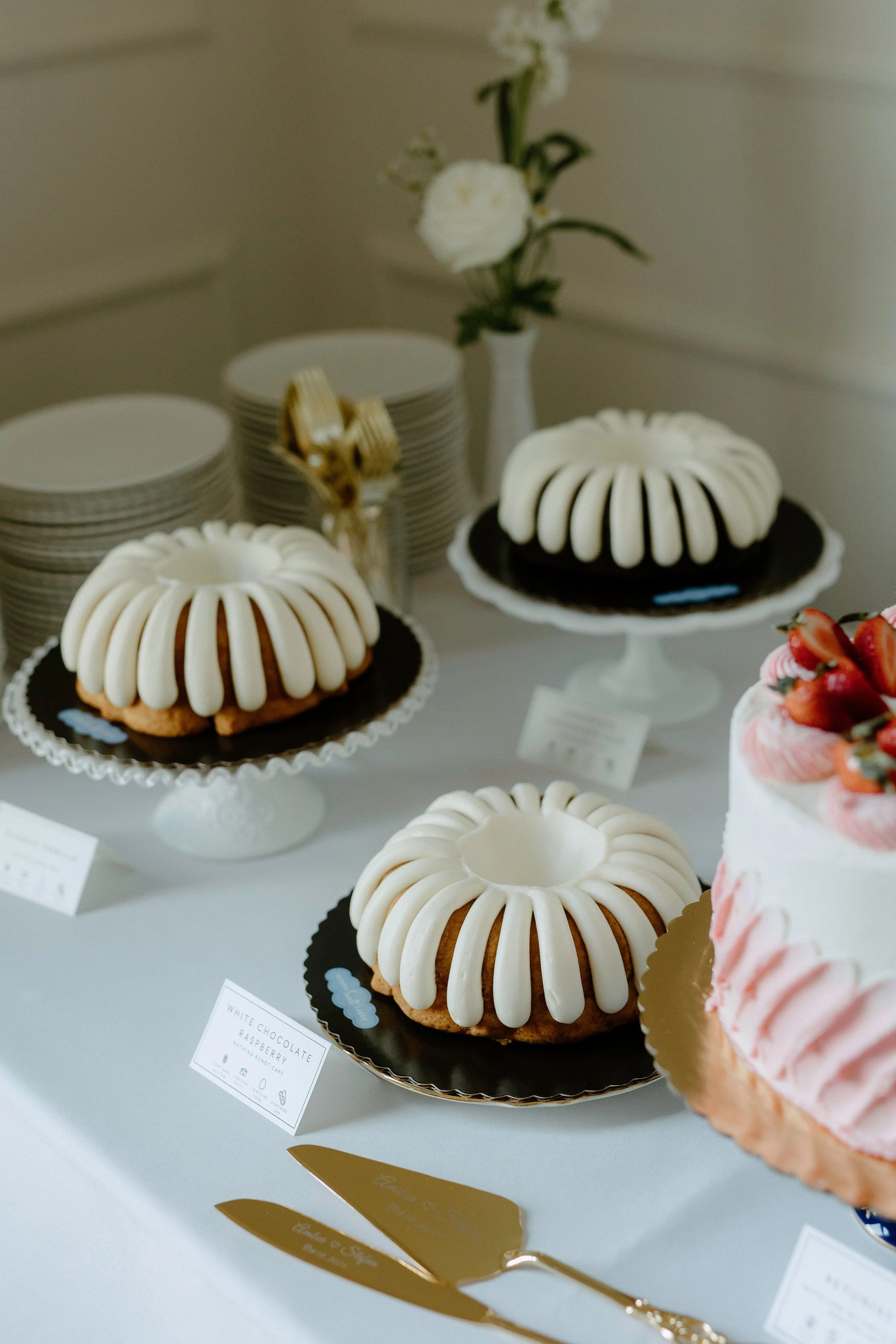 Assorted bundt cakes displayed on vintage milk glass cake stands with custom food placards and allergen designations — a thoughtful, heirloom-inspired dessert setup by Beam & Bramble