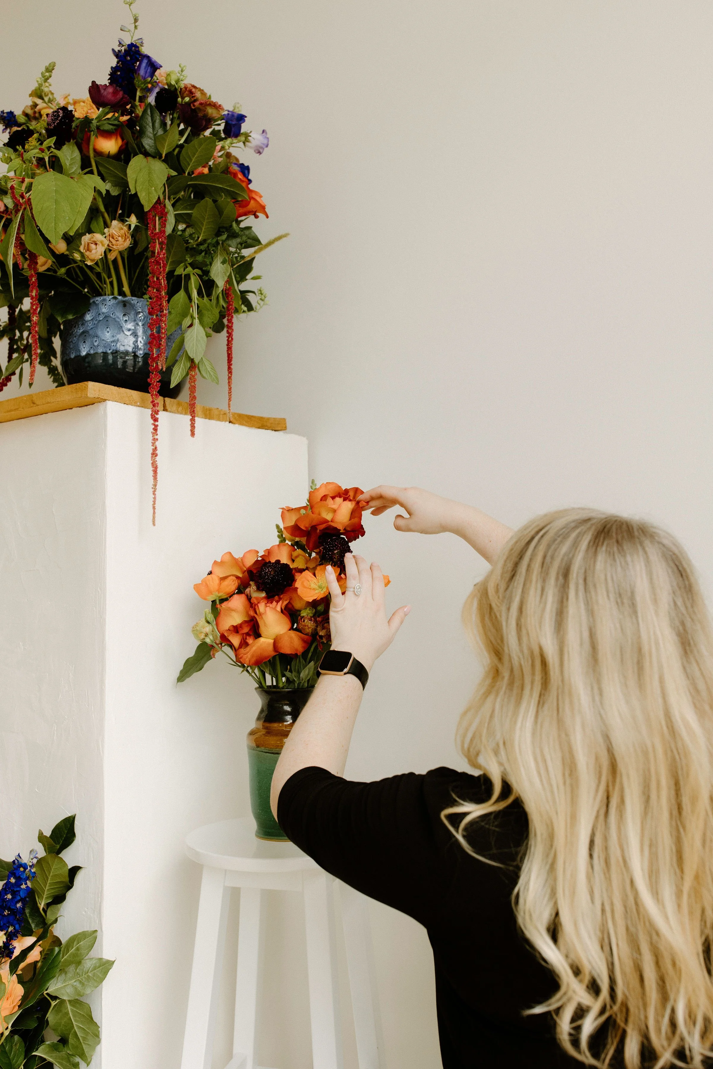 Founder styling a petite floral arrangement on a white stool, with rich jewel-toned florals and trailing greenery placed beneath a curated floral shelf install.