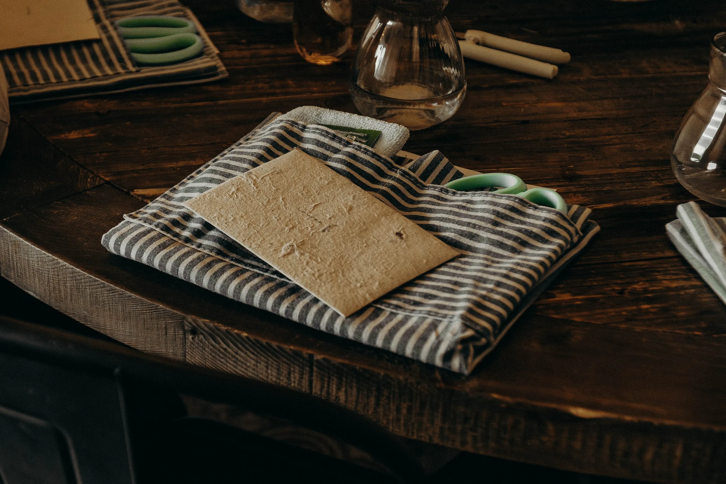 A wooden table with a folded striped cloth, a pair of green-handled scissors, a piece of parchment paper, and glass vases with water.