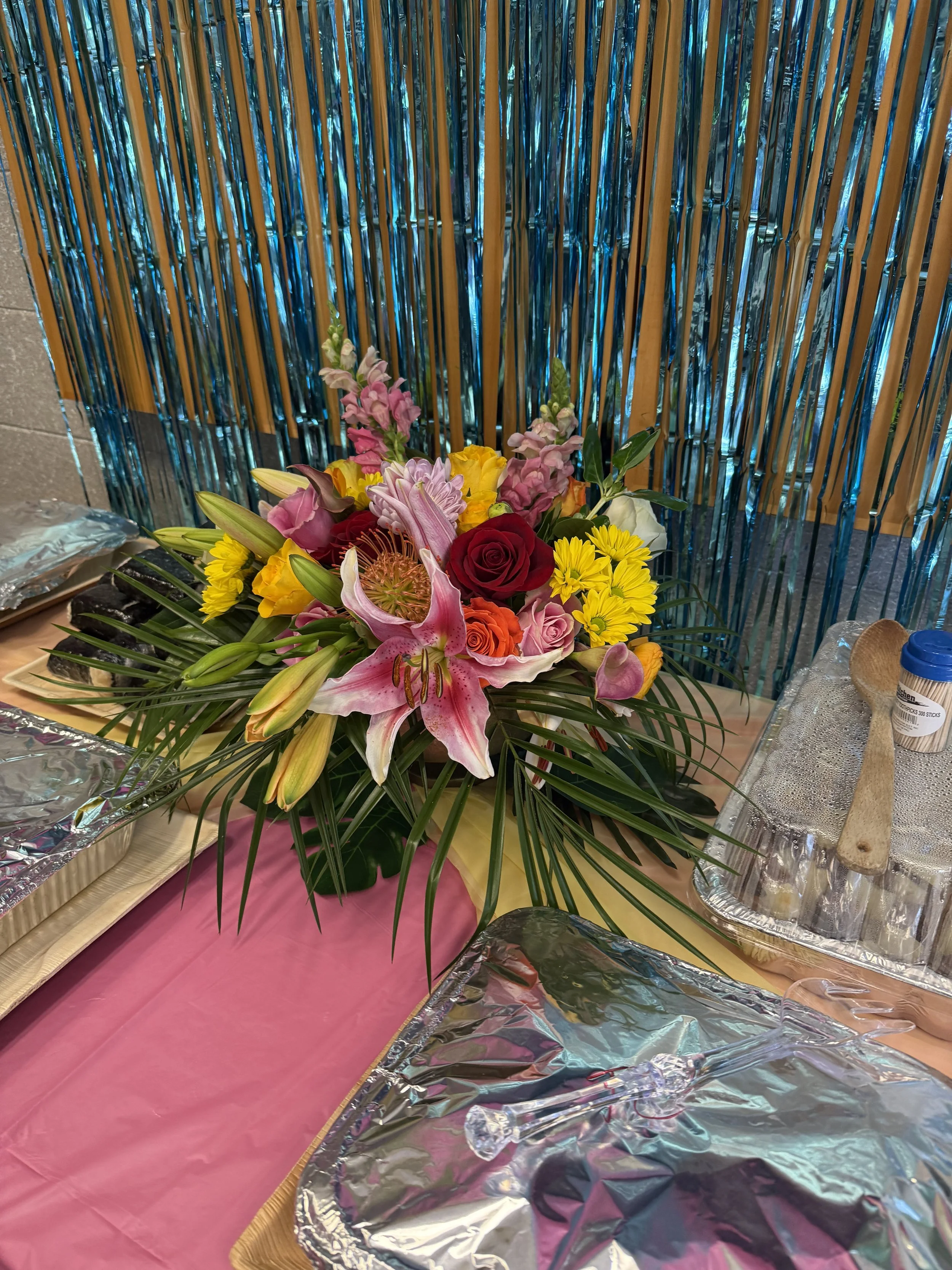 Vibrant tropical floral centerpiece with lilies, palm fronds, and colorful ribbons displayed in front of a teal and gold fringe backdrop