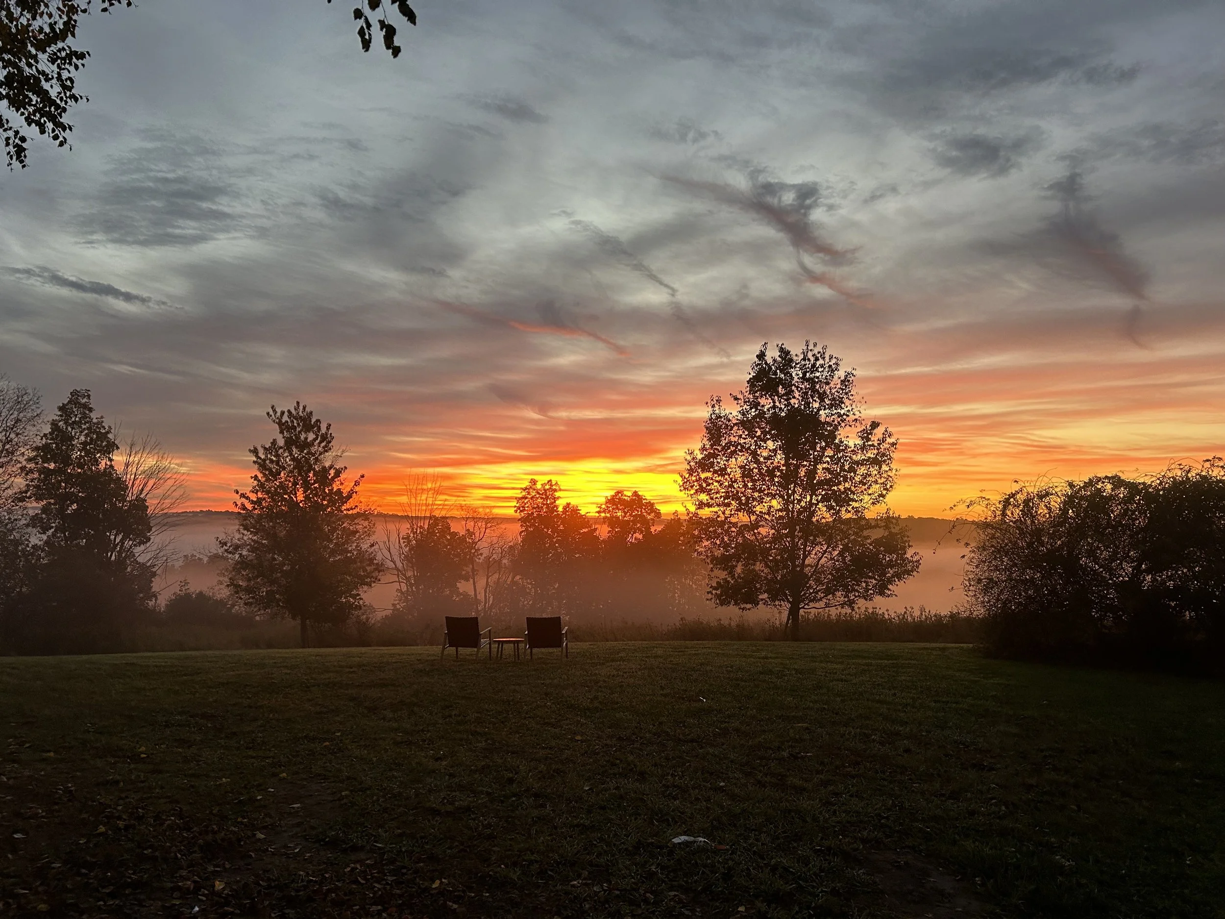 A tranquil sunset landscape with colorful sky, silhouetted trees, and two chairs facing the horizon on a grassy field.