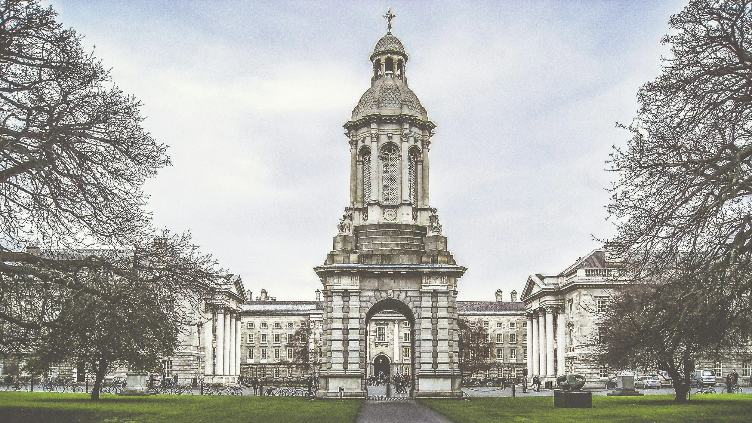 A historic building with a tall clock tower featuring a cross at the top, surrounded by leafless trees and green grass, on a cloudy day.