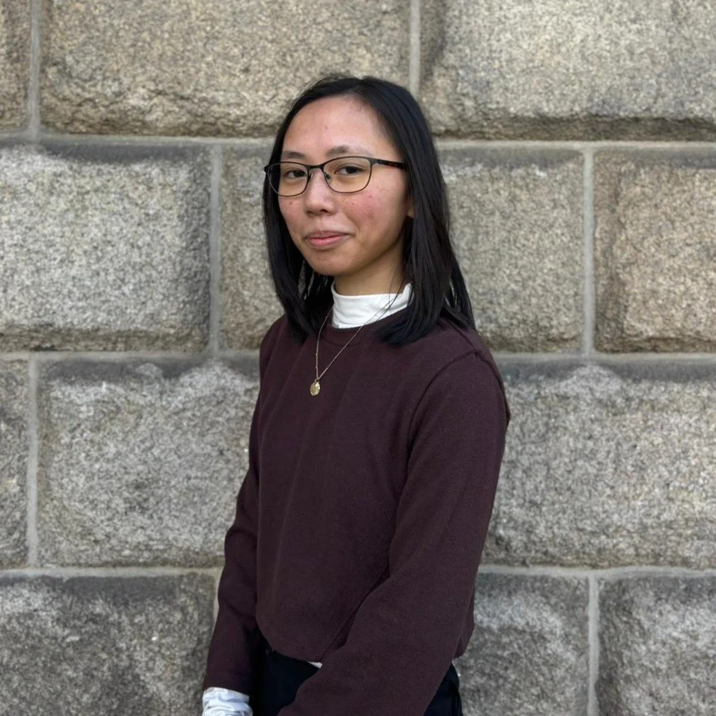 A woman with glasses and shoulder-length black hair posing in front of a stone wall, wearing a maroon sweater over a white shirt and a gold necklace.
