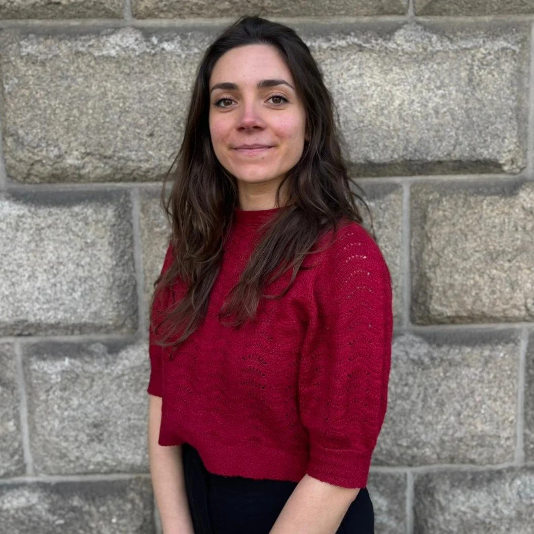 A young woman with long dark hair, wearing a red sweater, standing in front of a stone wall and looking at the camera with a slight smile.