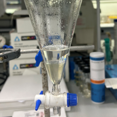 A laboratory setup with a glass funnel connected to a tube with a blue valve, containing a clear liquid, with lab equipment and containers in the background.