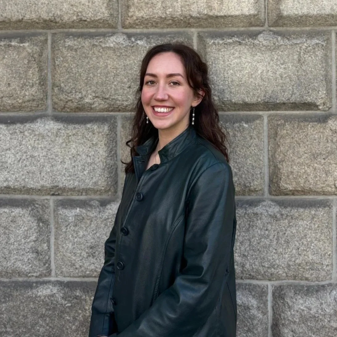 A young woman with dark brown wavy hair, smiling, standing against a gray stone wall, wearing a dark leather jacket and dangling earrings.