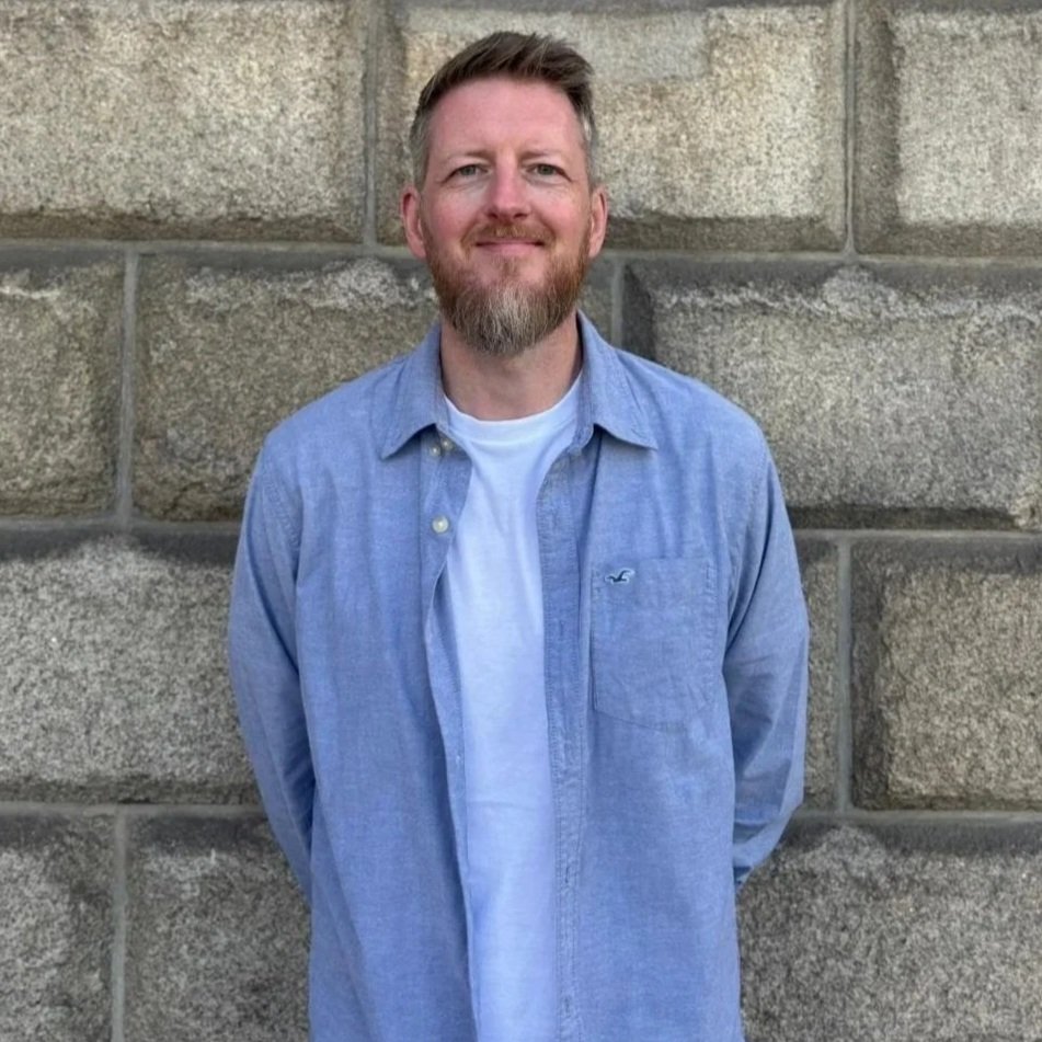 A man with a beard and short hair standing in front of a stone wall, smiling, wearing a blue button-up shirt over a white t-shirt.