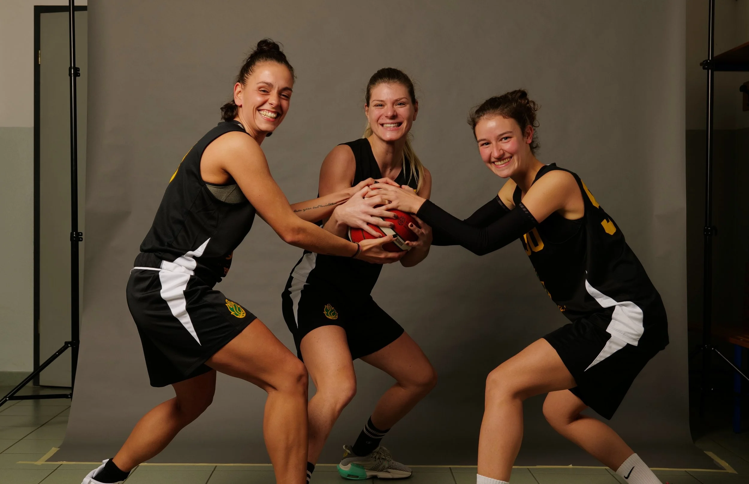 Tre giocatrici di basket della promozione femminile della pallacanestro ponzano sorridenti in posa con una palla, indossano divise sportive nere con dettagli bianchi e gialli, durante una sessione fotografica in uno studio con sfondo grigio.
