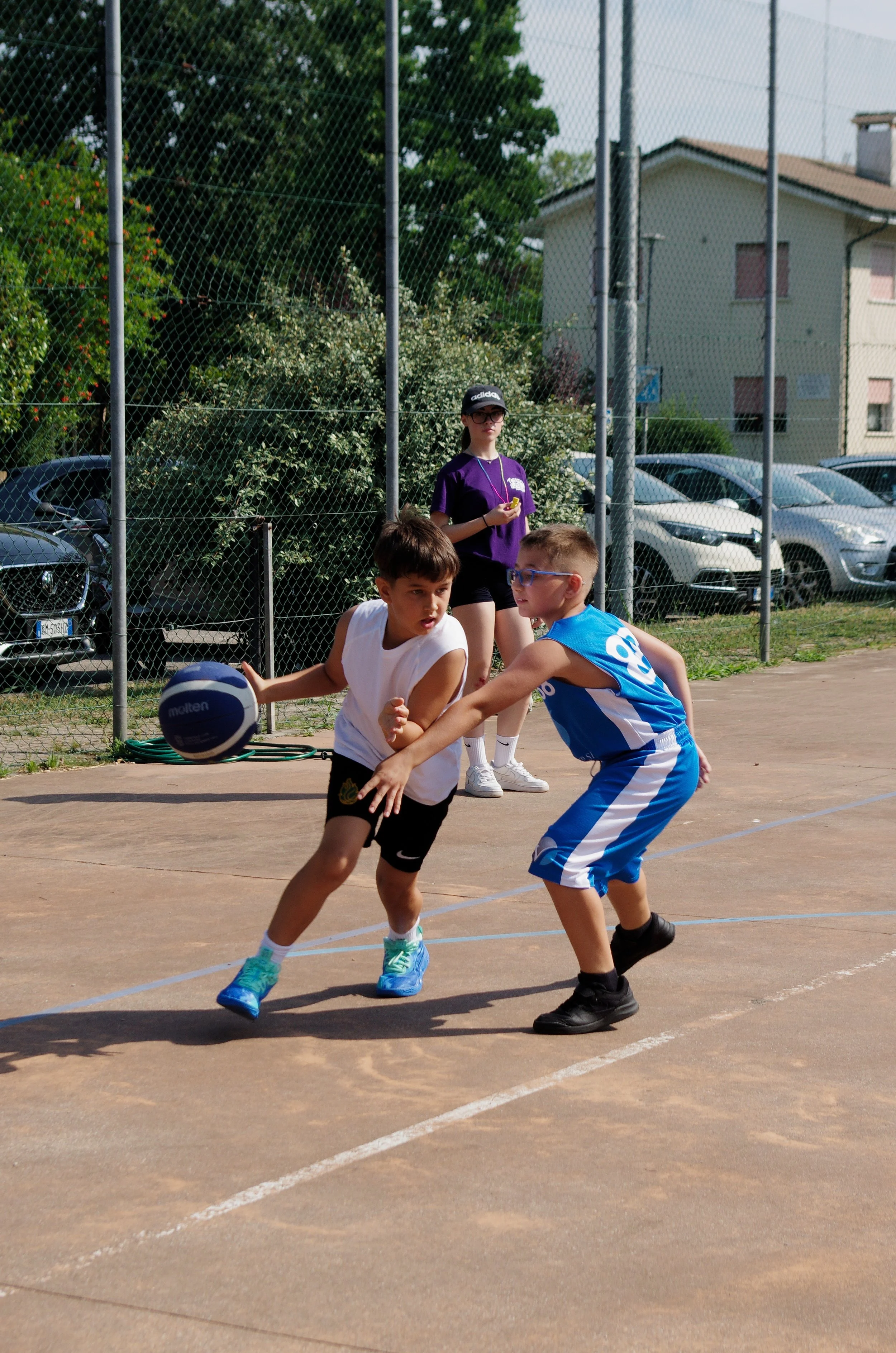 Due bambini giocano a pallacanestro su un campo all'aperto, con una ragazza in piedi sullo sfondo e auto parcheggiate dietro una rete.