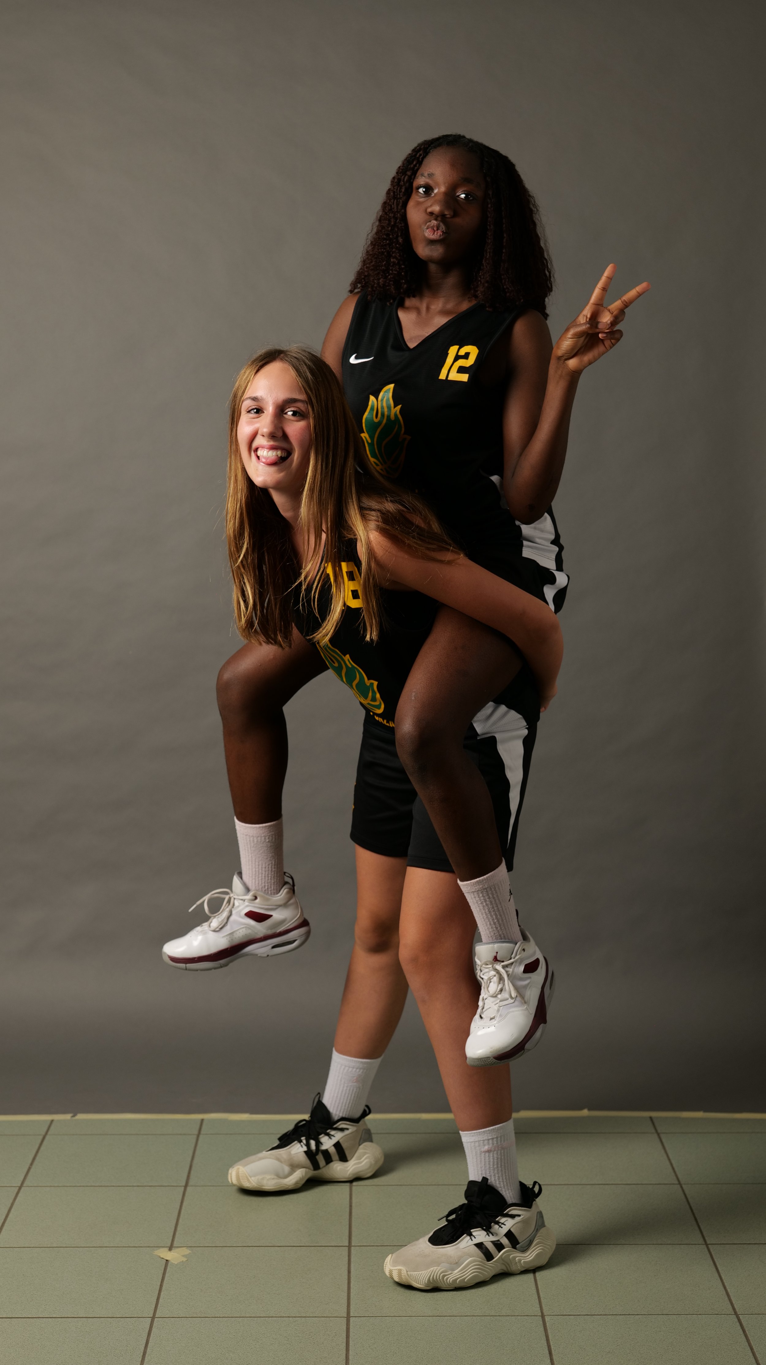 Due atlete under 14 femminile di basket della pallacanestro ponzano in tenuta sportiva nera, una con maglietta numero 12 e l'altra numero 8, si divertono in uno scambio giocoso, con una ragazza che fa una V e un'espressione sorridente.