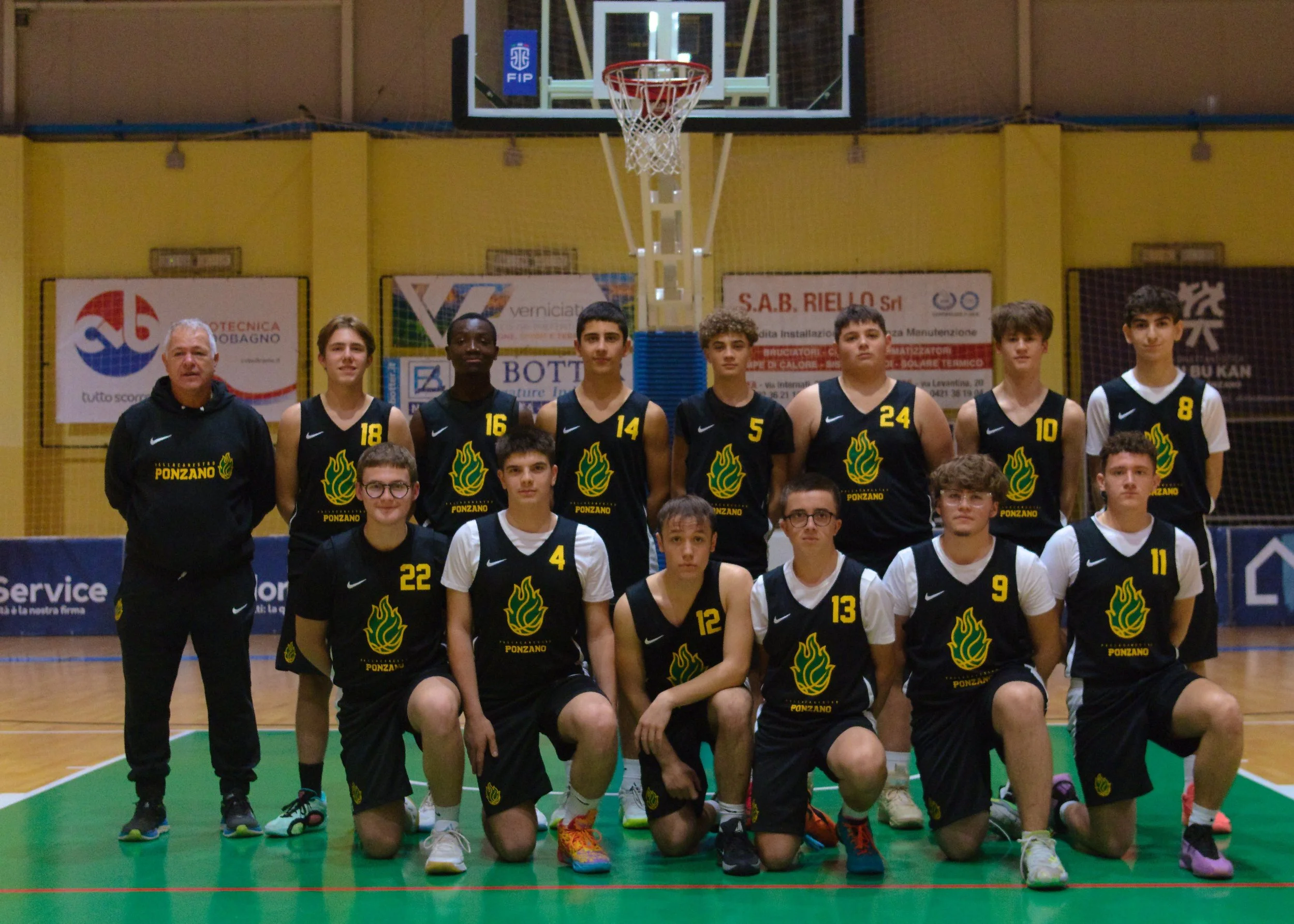 Gruppo di giovani giocatori di basket under 17 maschile e l'allenatore Paolo De Rossi posano per la foto di squadra con divise nere con logo della pallacanestro ponzano e numeri gialli, in piedi davanti a un canestro da basket.