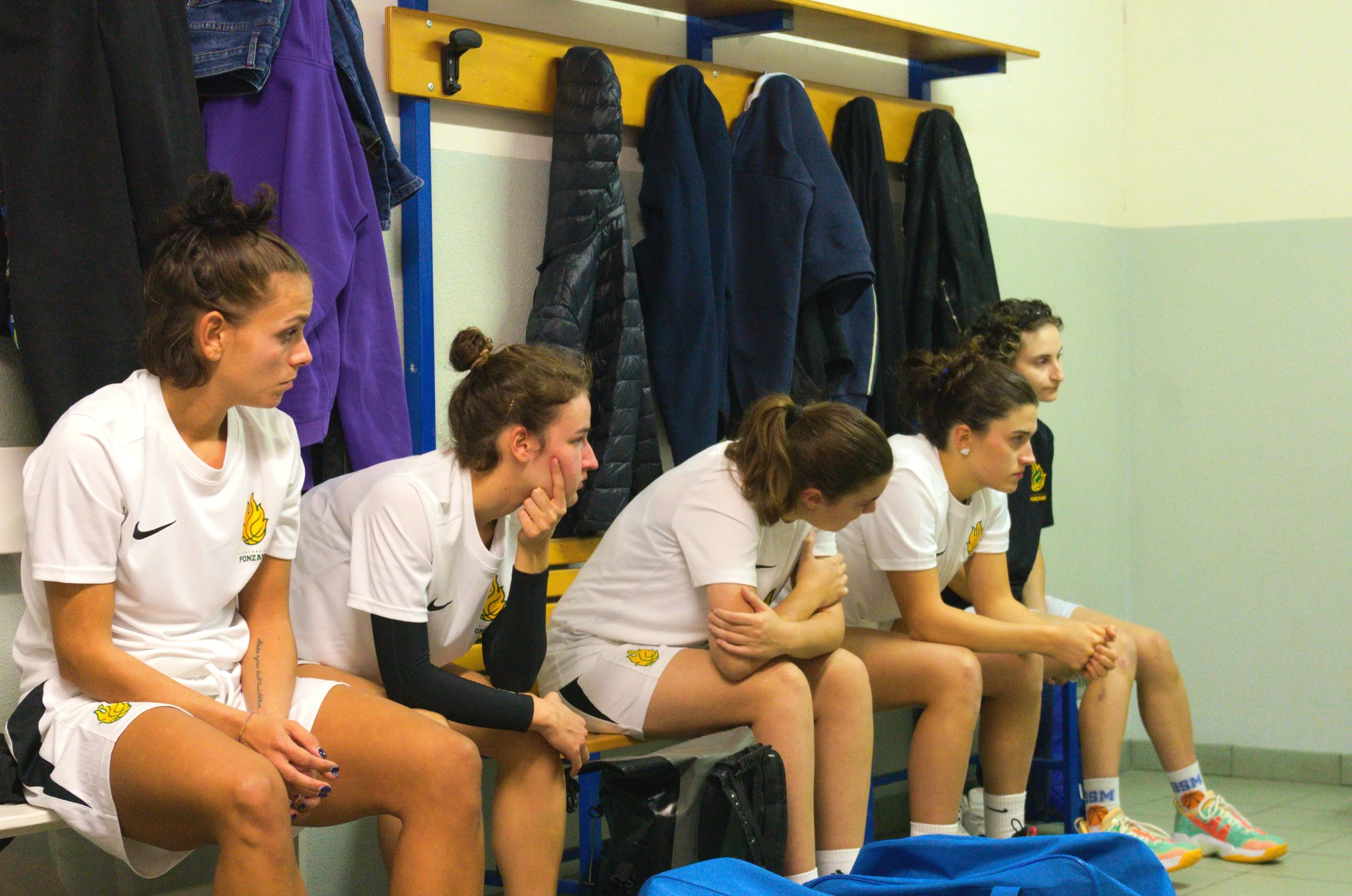 Cinque giocatrici della promo femminile sedute su una panchina in spogliatoio, indossando divise da basket bianche con il logo della pallacanestro ponzano, con giacche appese dietro di loro.