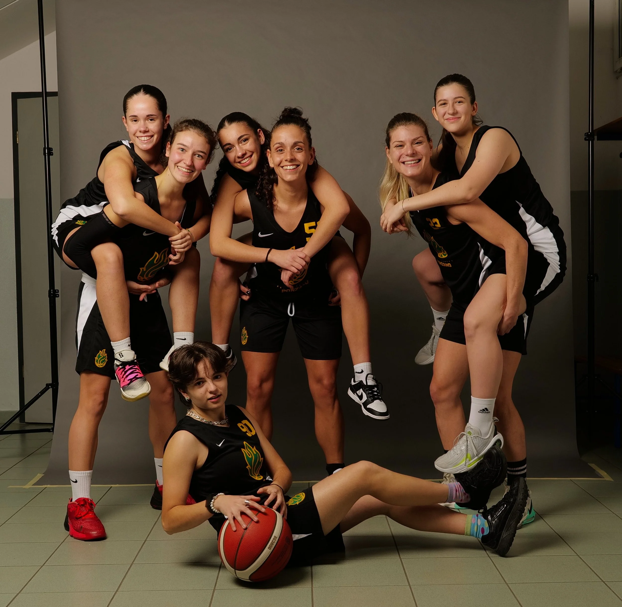 Gruppo di giovani atlete della promozione femminile di pallacanestro ponzano in divisa da basket nera sorridenti, alcune in pose di gioco, una seduta con palla, in uno studio con sfondo grigio.