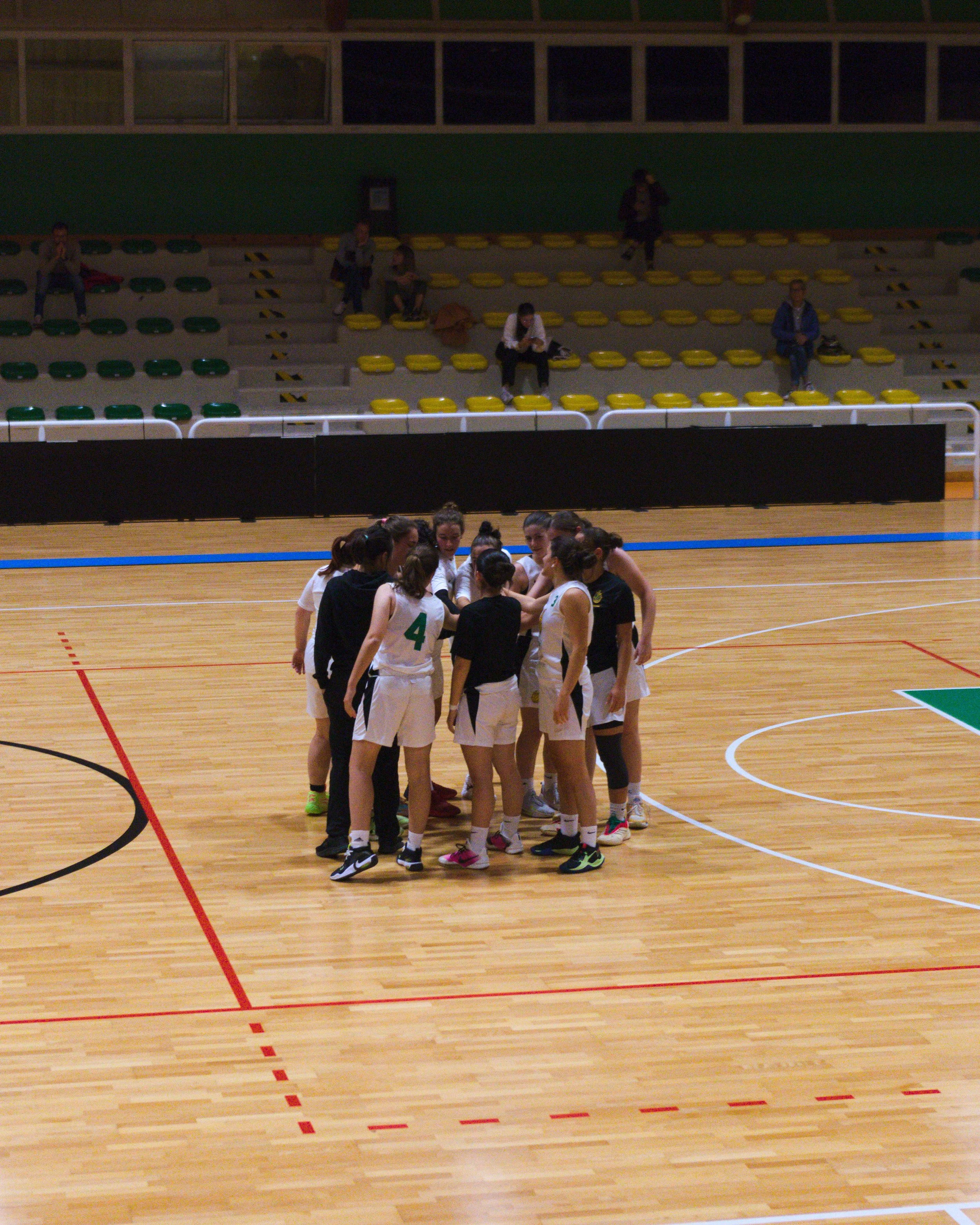 Gruppo di giocatrici di pallacanestro Ponzano che si riuniscono in cerchio a inizio partita nel palazzetto Palacicogna, con alcune persone sedute sulle tribune sullo sfondo.