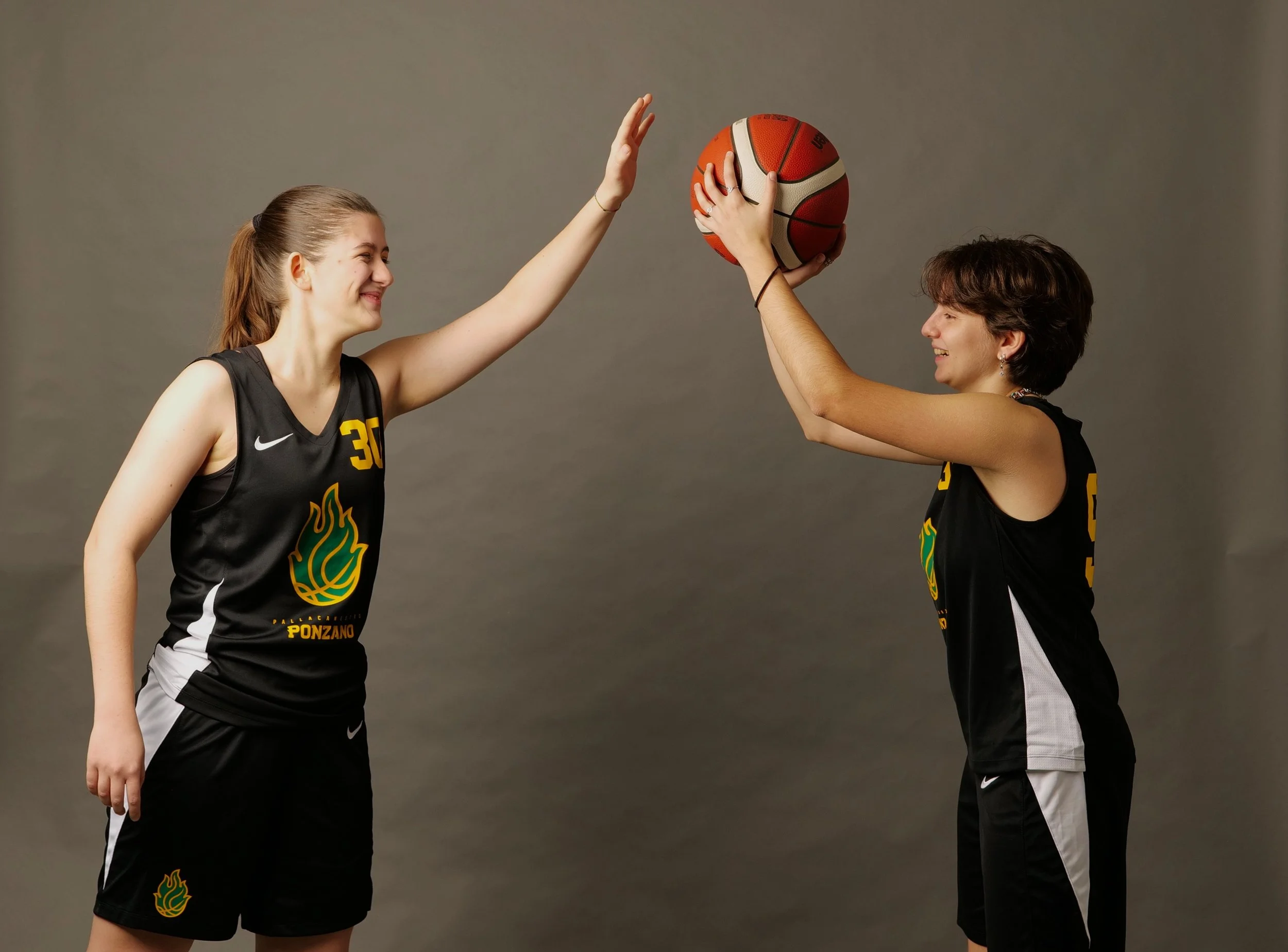 Due donne della promo femminile  della pallacanestro ponzano in uniforme da basket, si scambiano un pallone da basket in un gesto di gioco amichevole, entrambe sorridenti su uno sfondo neutro.